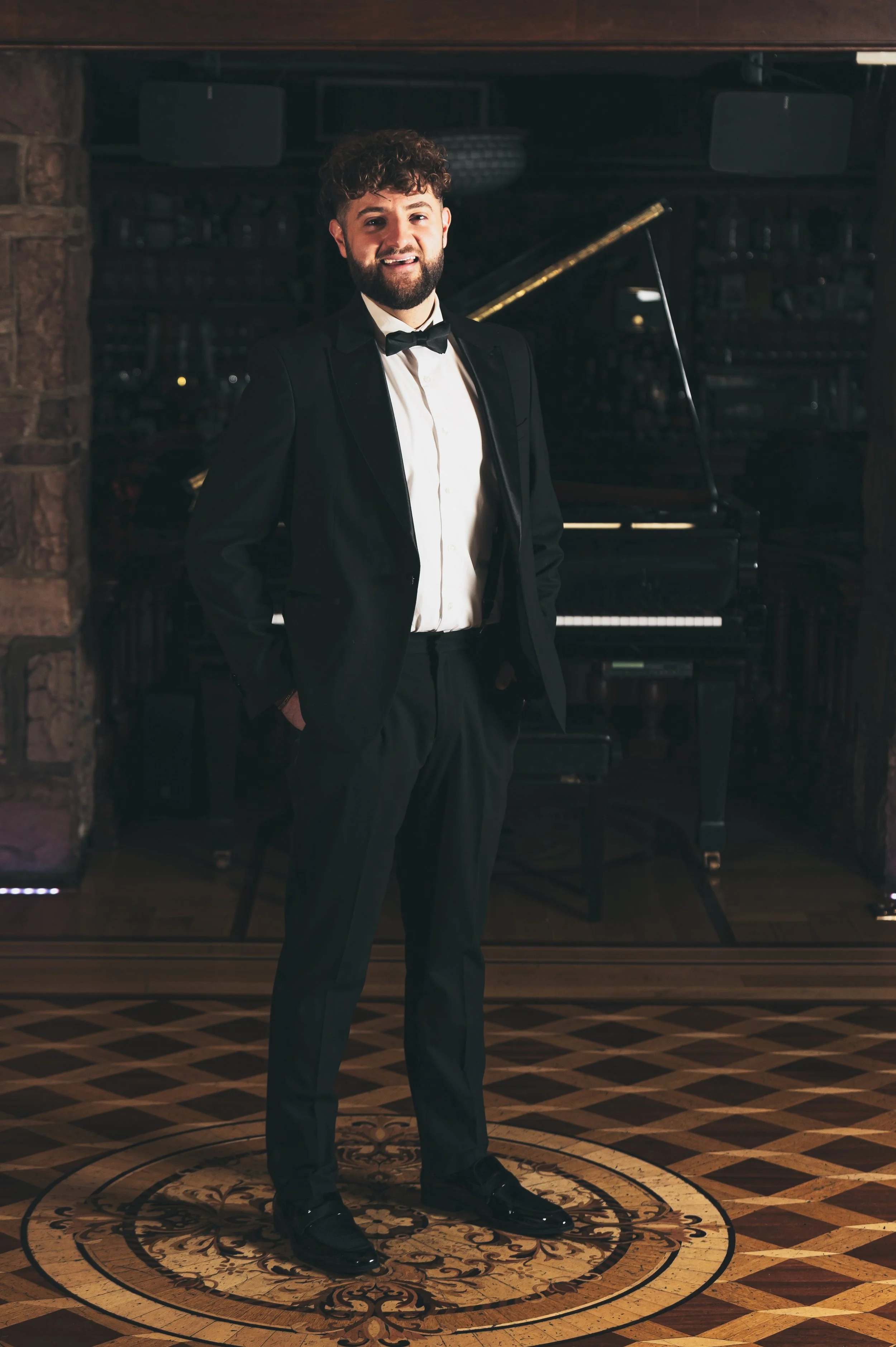 A man in a black tuxedo with a bow tie stands on a decorative wooden floor in front of a black grand piano, smiling at the camera.
