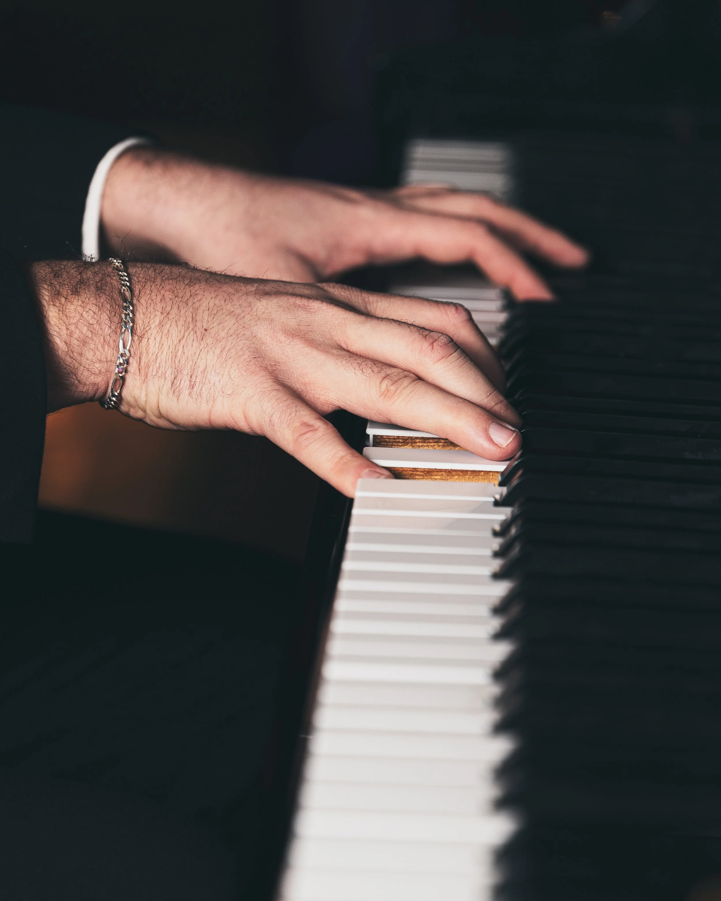 Person playing piano, focusing on hands pressing keys, wearing a bracelet and shirt cuffs.