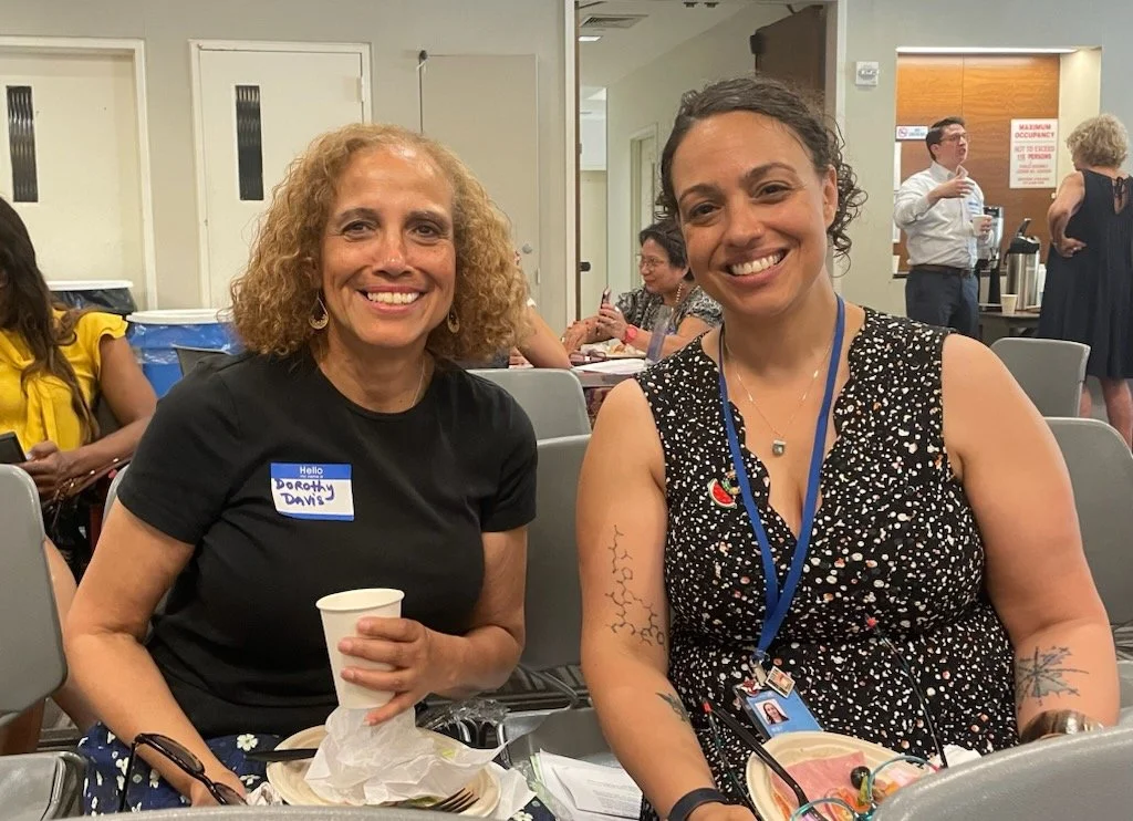 Two women sitting at a table in a conference room smiling at the camera. They have name tags, drinks, and food in front of them. There are other people in the background, some standing and some seated.