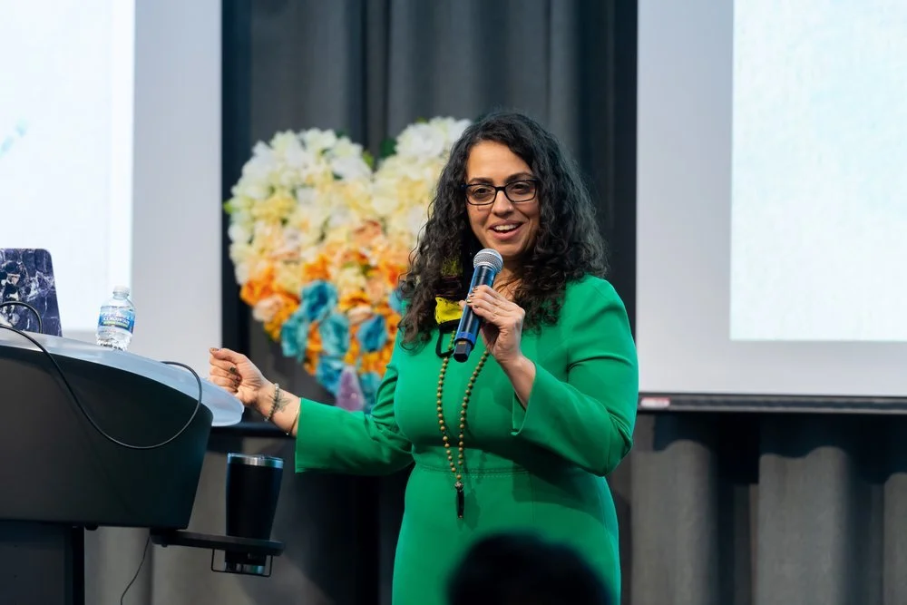 A woman in a green dress holding a microphone, giving a presentation in front of a large floral arrangement and a projection screen.