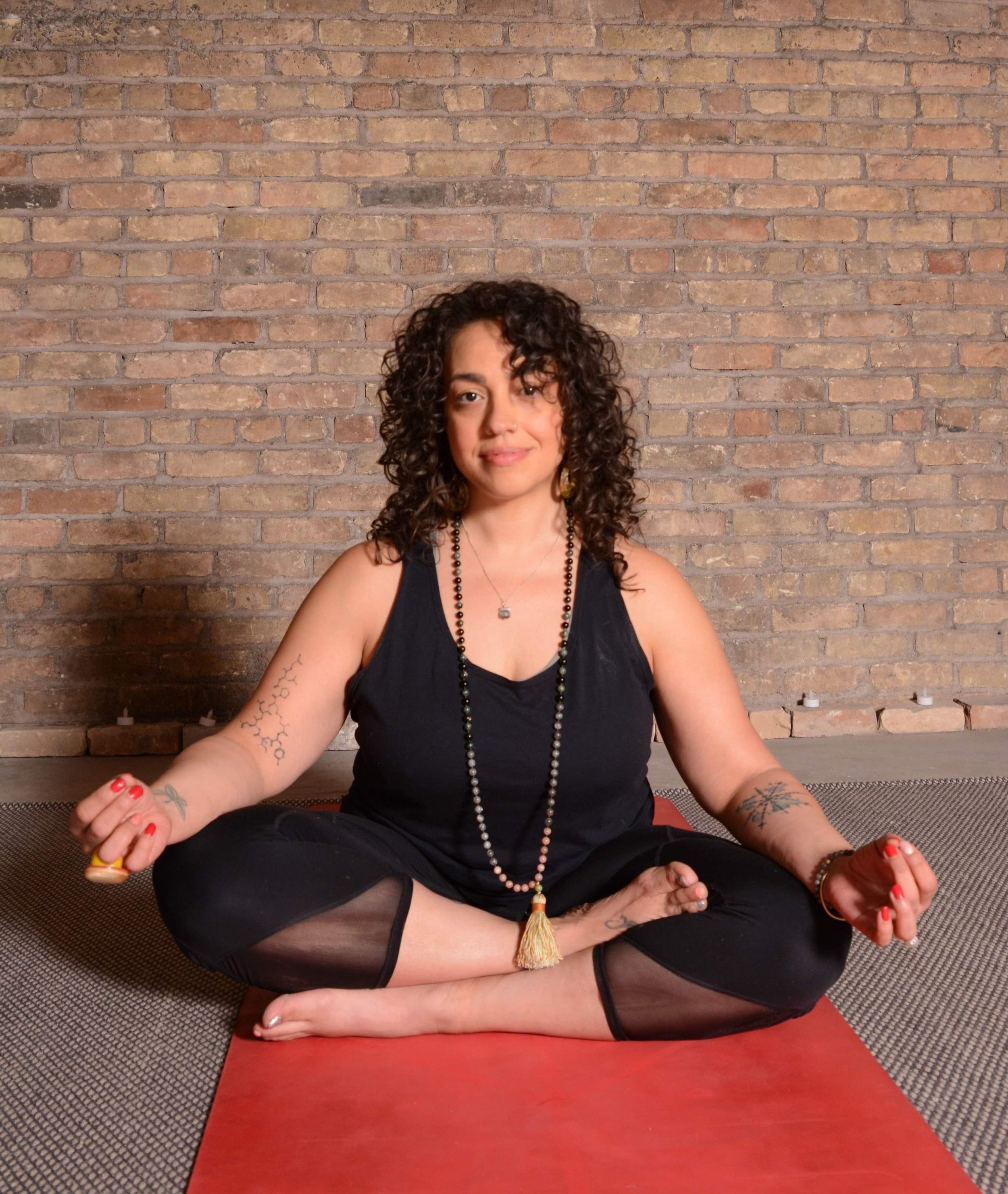Woman with curly hair practicing yoga in a seated meditation pose on a red mat, against a brick wall background.