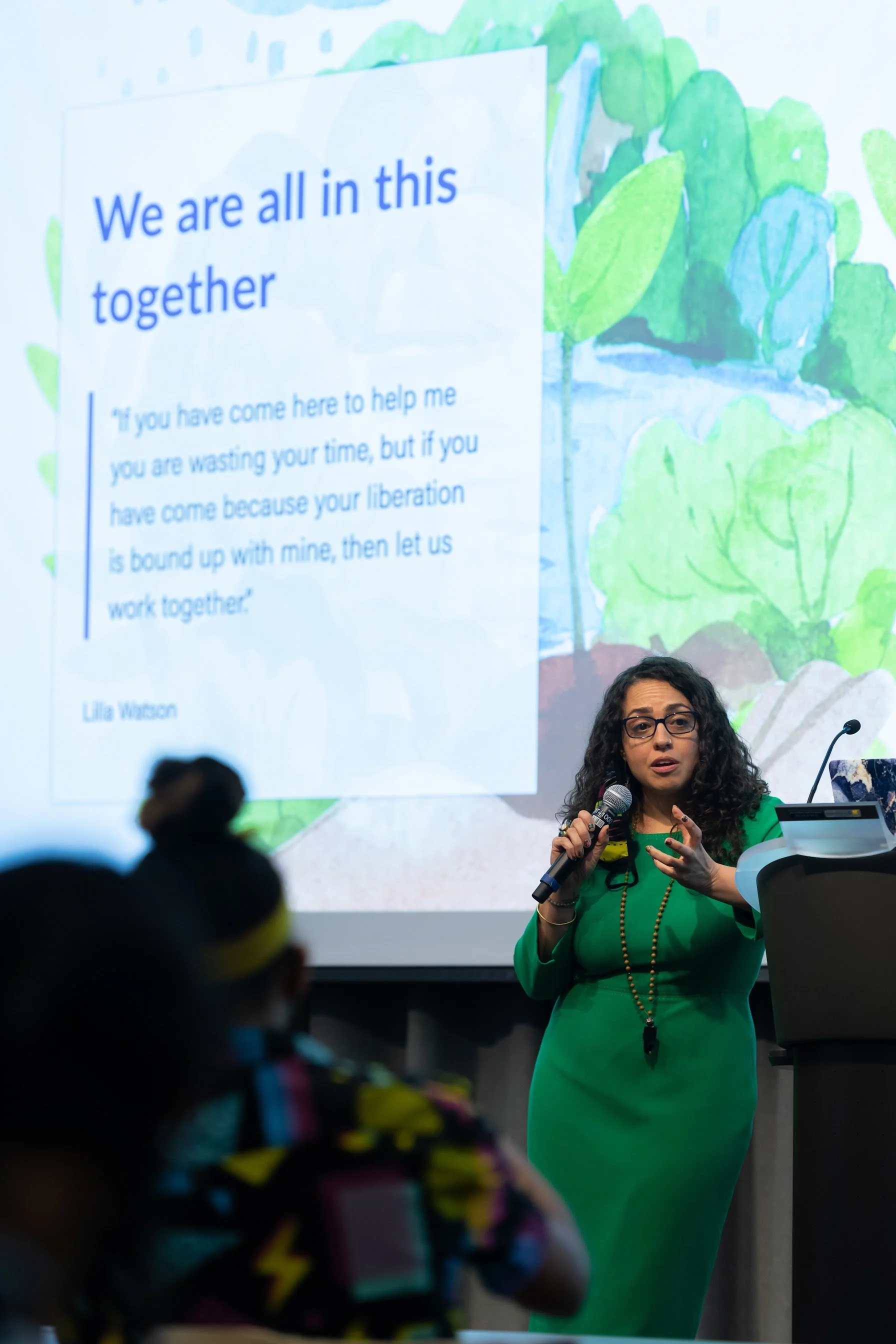 A woman in a green dress holding a microphone in front of a large projected slide that features a quote by Lila Watson, with a background of colorful watercolor leaves and flowers.