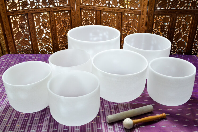 Seven white crystal singing bowls of various sizes arranged on a purple cloth with a decorative wooden screen background, along with a mallet and striker for playing the bowls.