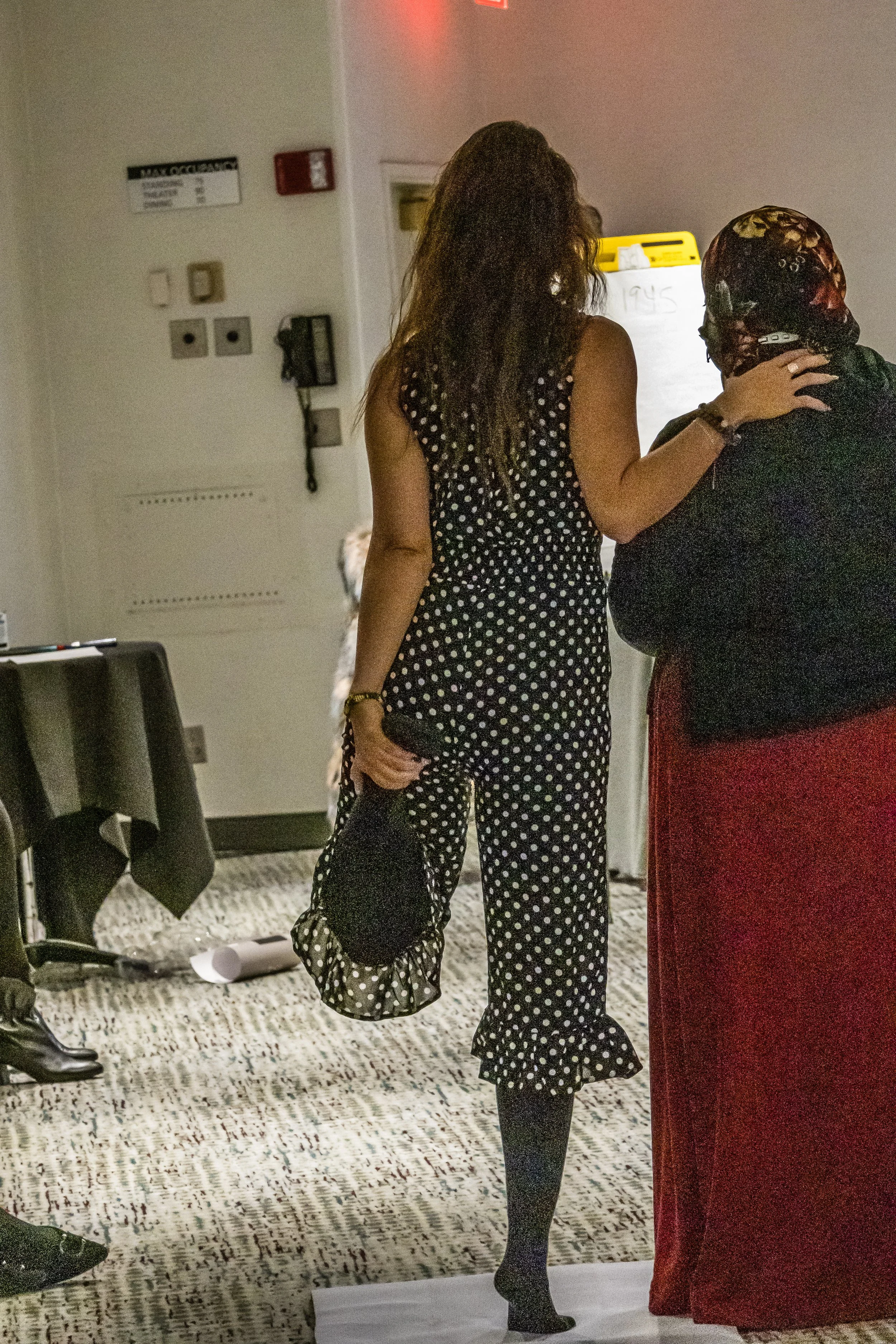Two women in conversation, one in a polka dot outfit holding a matching polka dot bag, and the other wearing a skull cap and black top, standing near a chalkboard or whiteboard in an indoor setting.