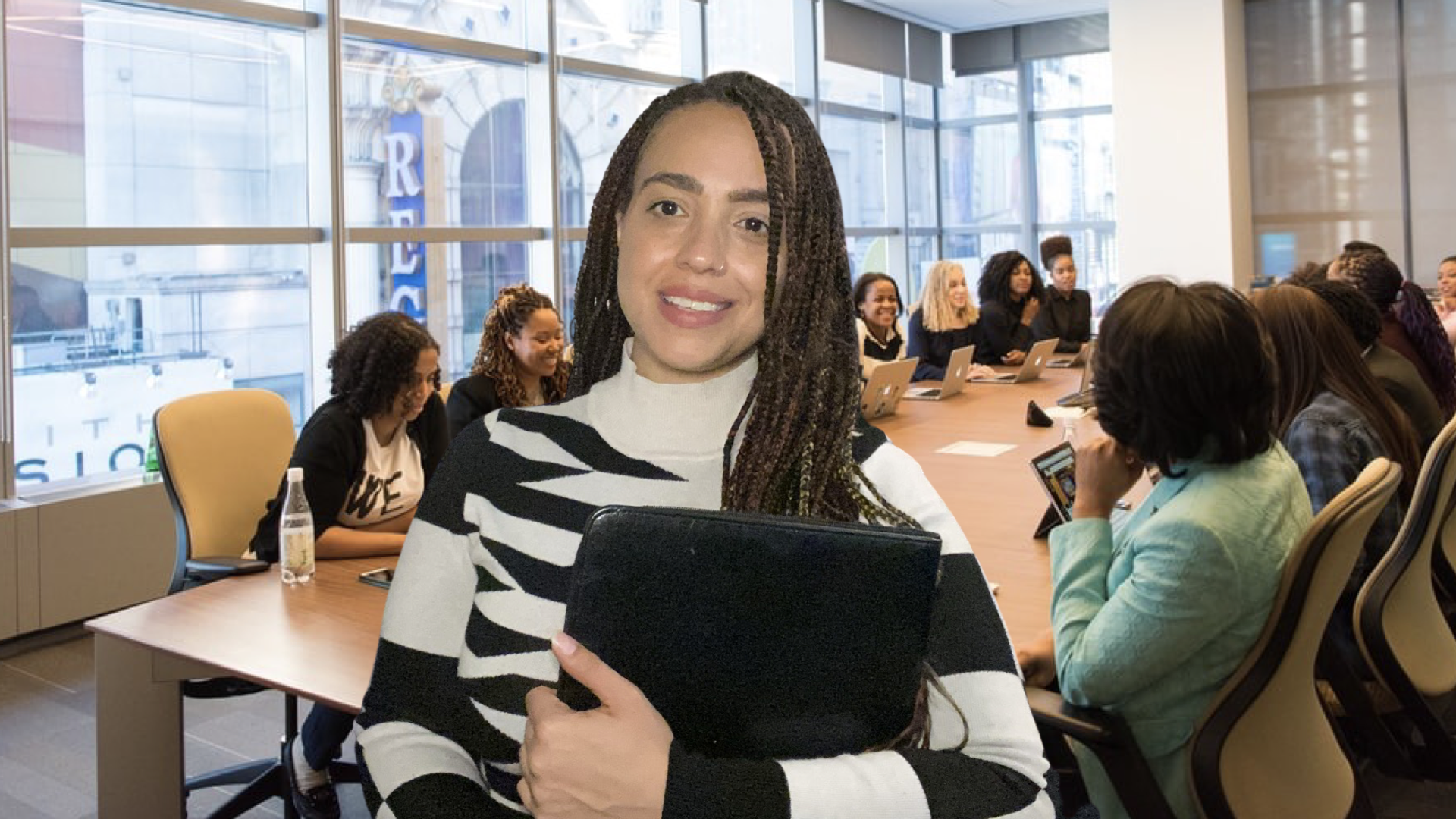 A woman with braided hair holding a folder and smiling in a conference room with a diverse group of women working on laptops at a long table with floor-to-ceiling windows behind them.