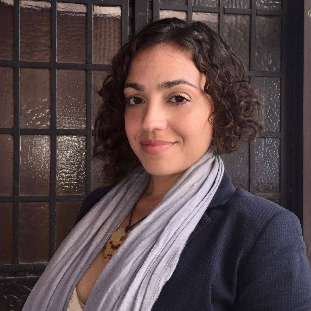 A woman (Rebecca Irby) with shoulder-length curly brown hair smiling at the camera, wearing a dark blazer and a light gray scarf, standing in front of a window with textured glass panes.