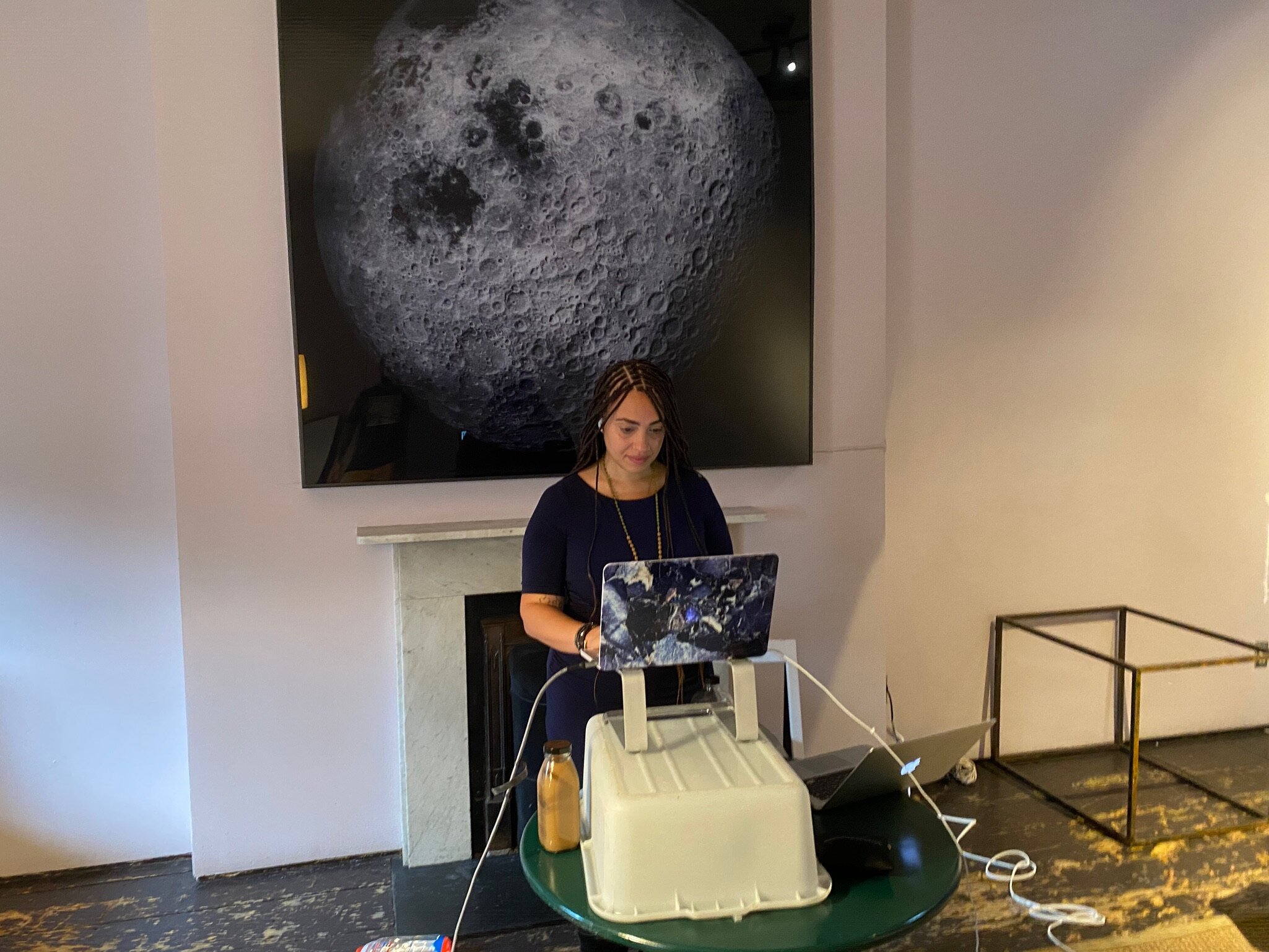 A woman (Rebecca Irby) with braided hair stands in front of a fireplace, working on a laptop with a galaxy-patterned cover, while a large screen behind her displays a detailed image of the moon.