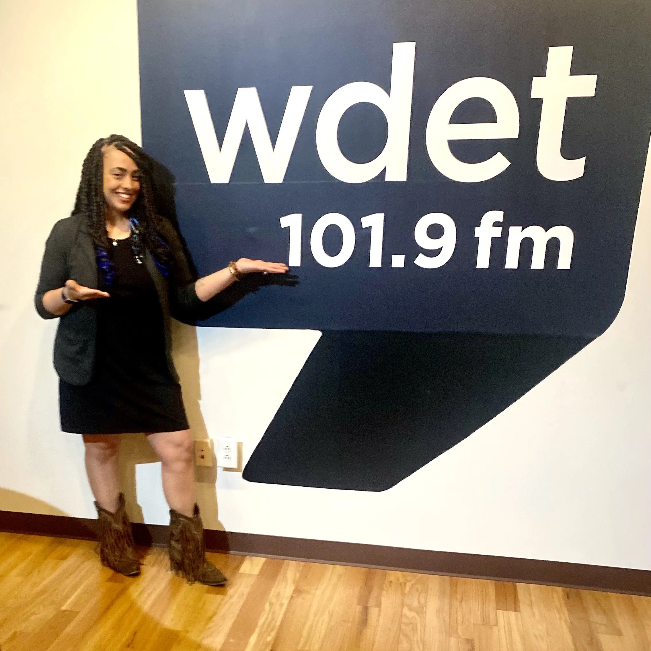 A woman (Rebecca Irby) standing next to a large wall sign displaying the logo and frequency of WDET 101.9 FM radio station, with her arms raised as if presenting the sign, smiling and wearing a black dress, black blazer, and cowboy boots.