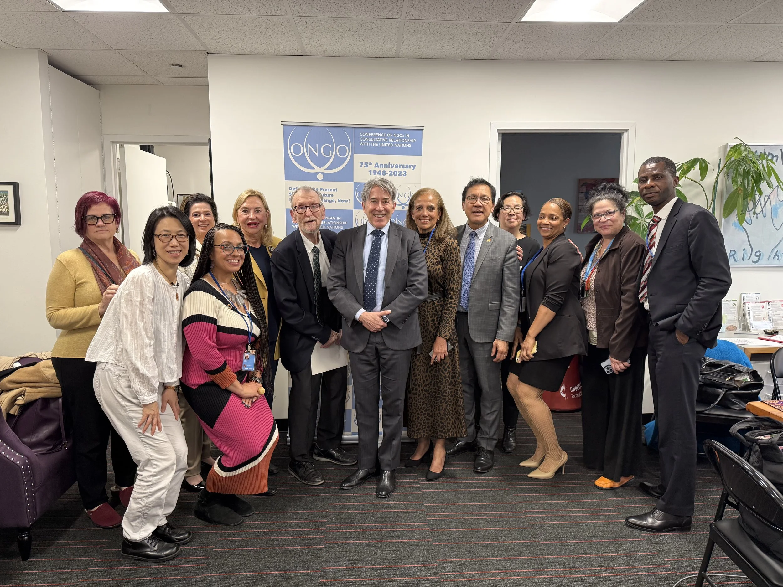 Group of diverse men and women posing for a photo in an office or conference room with a banner in the background celebrating the 75th anniversary of an NGO. Everyone is dressed in formal and semi-formal attire, smiling at the camera.