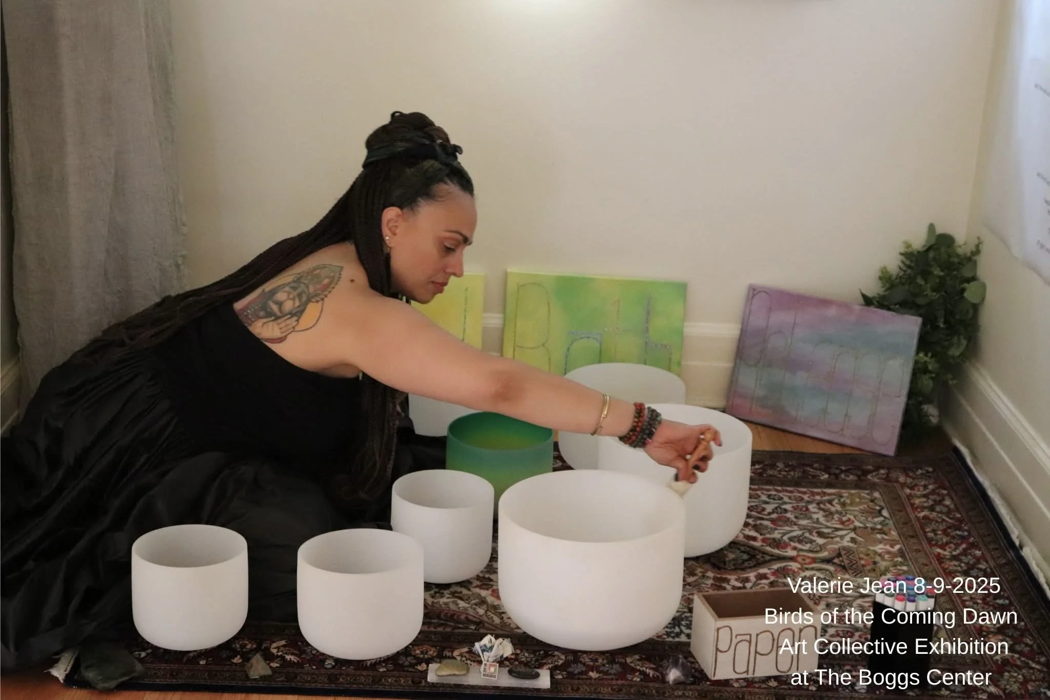 A woman with long dreadlocks, wearing a black dress, is sitting on a patterned rug playing crystal singing bowls of various sizes. There are colorful paintings behind her, and a small box labeled 'Pap' is on the floor.