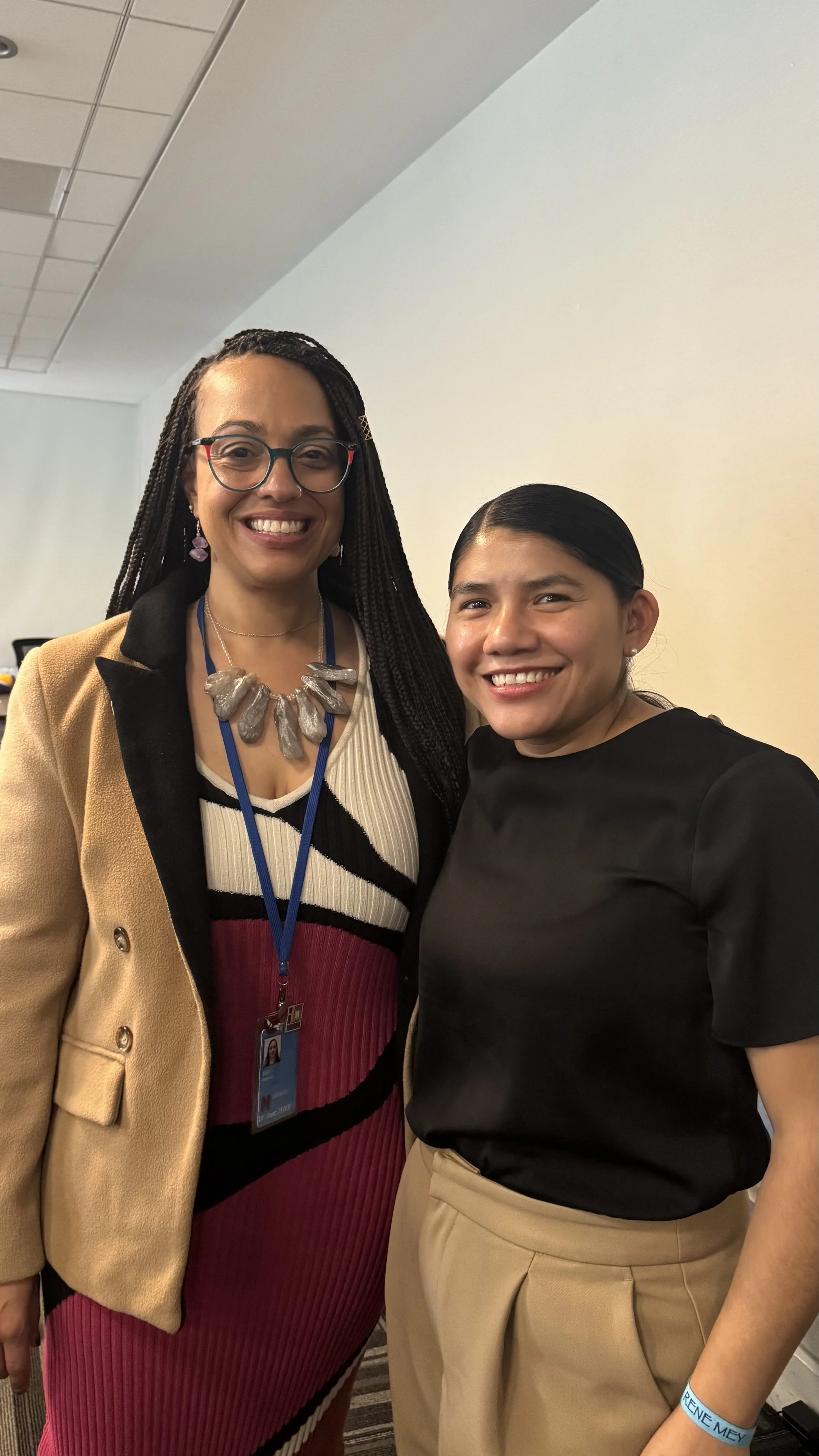 Two women smiling and standing close together in an indoor setting, one wearing glasses, a beige blazer, and a colorful dress, and the other in a black top and beige pants.