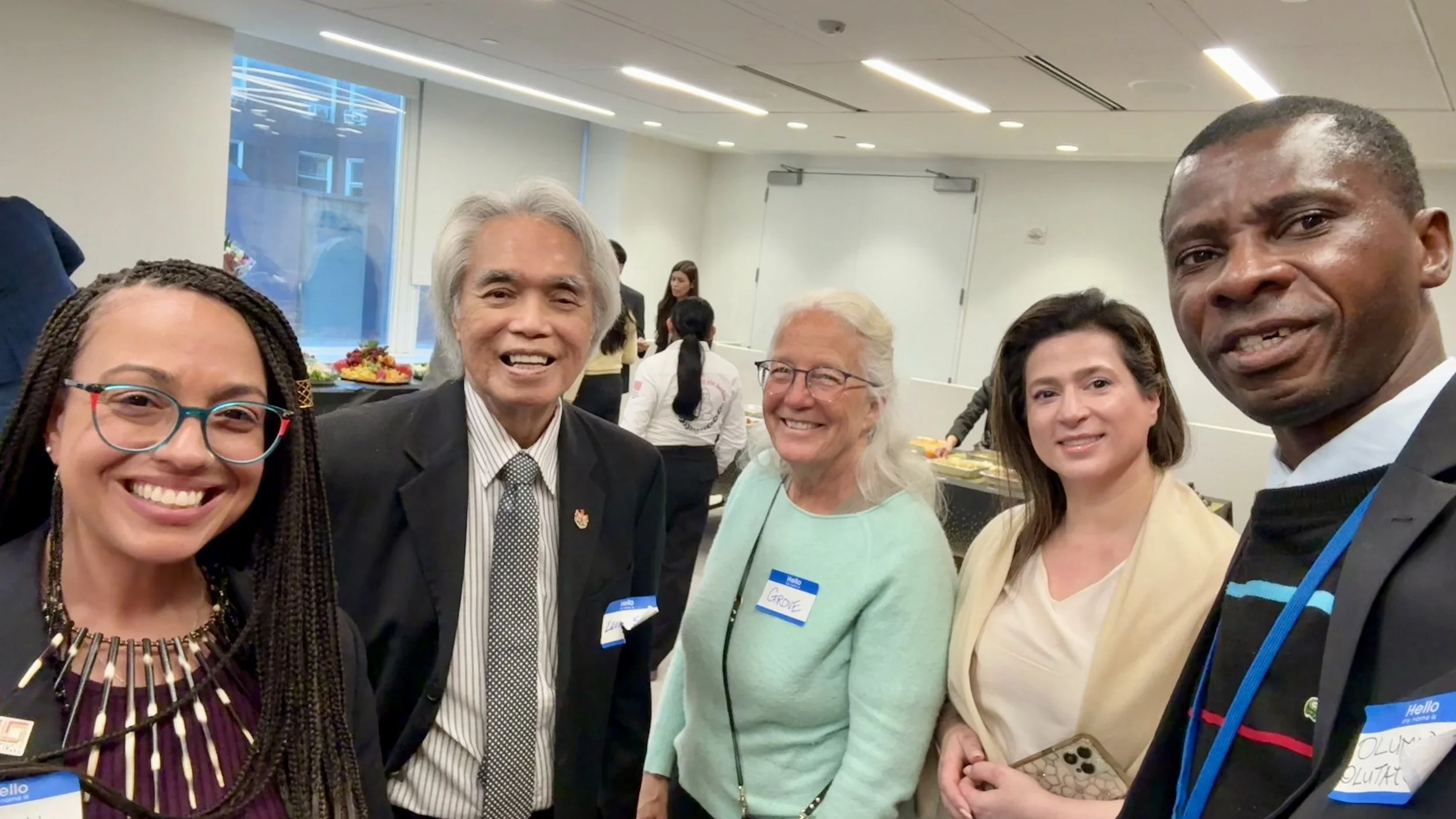 A group of five diverse people at a social gathering, smiling, with a buffet table in the background in a bright, modern indoor space.