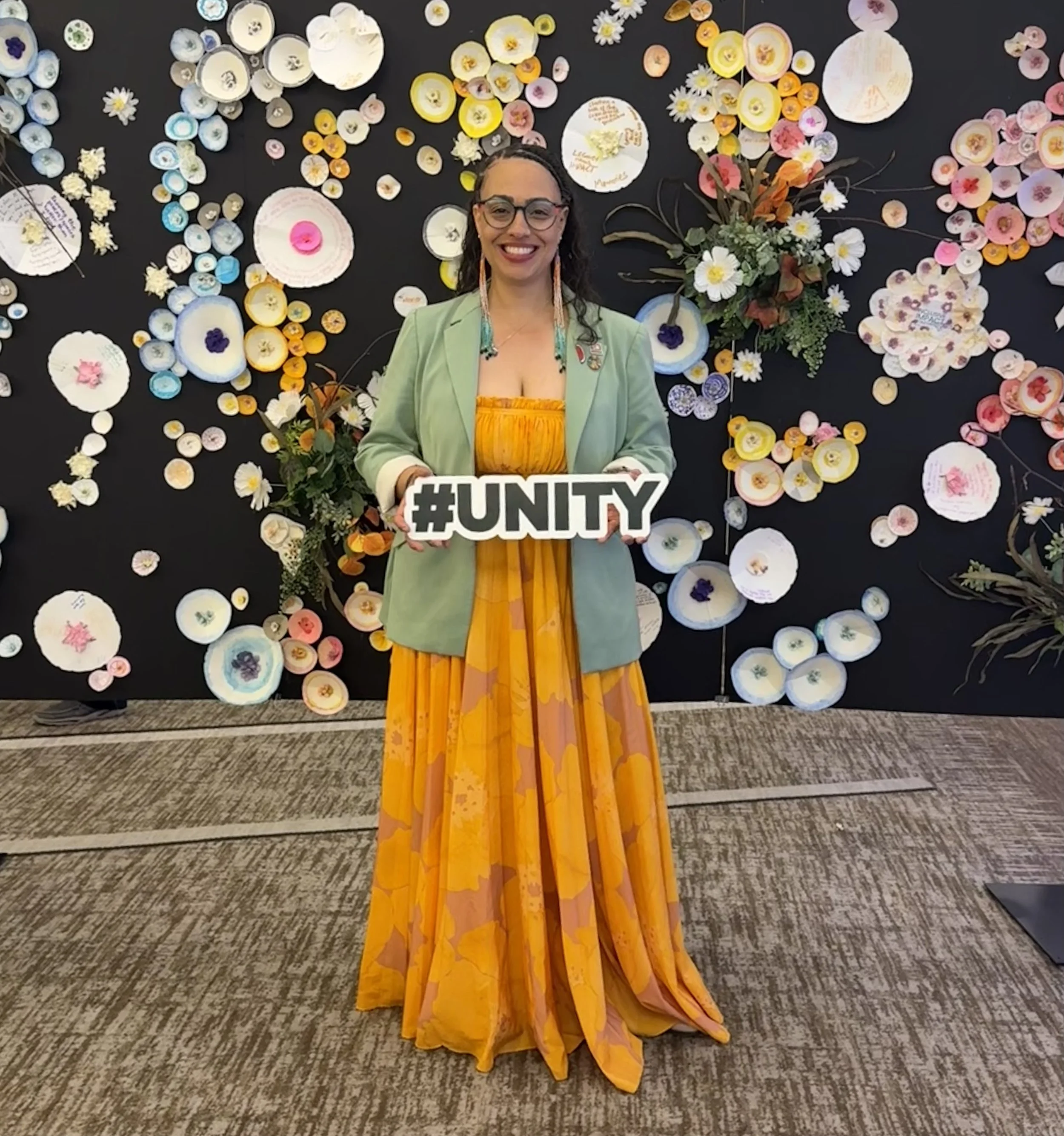 A woman standing in front of a floral wall holding a sign that reads '#UNITY.' She is wearing glasses, a green blazer, a yellow dress, and colorful earrings.