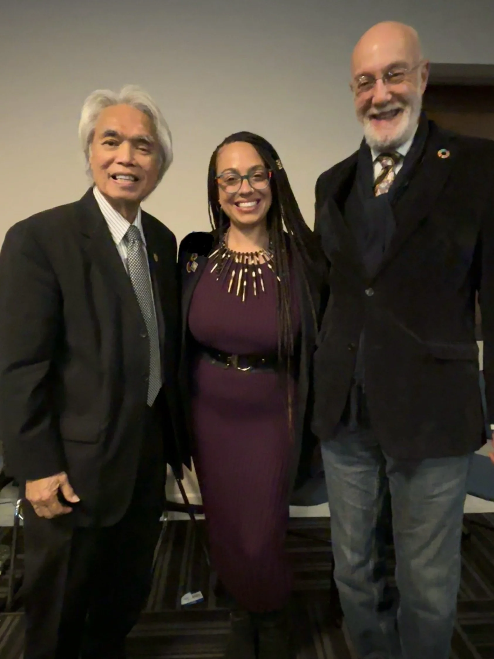 Three people smiling and standing together indoors, dressed in formal attire.