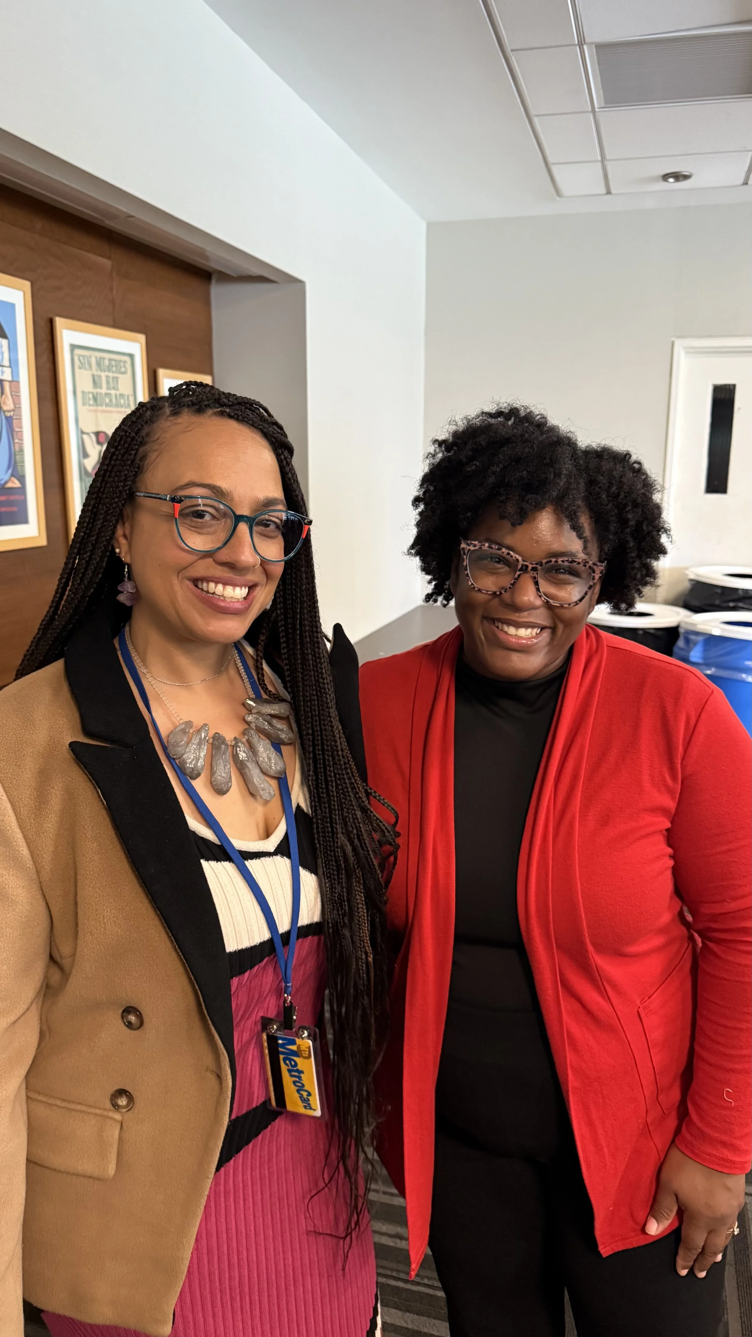 Two women smiling and standing close together, one wearing a tan blazer, colorful dress, and large necklace, the other in a red jacket and black top, with a background of recycling bins and framed artwork on a wall.