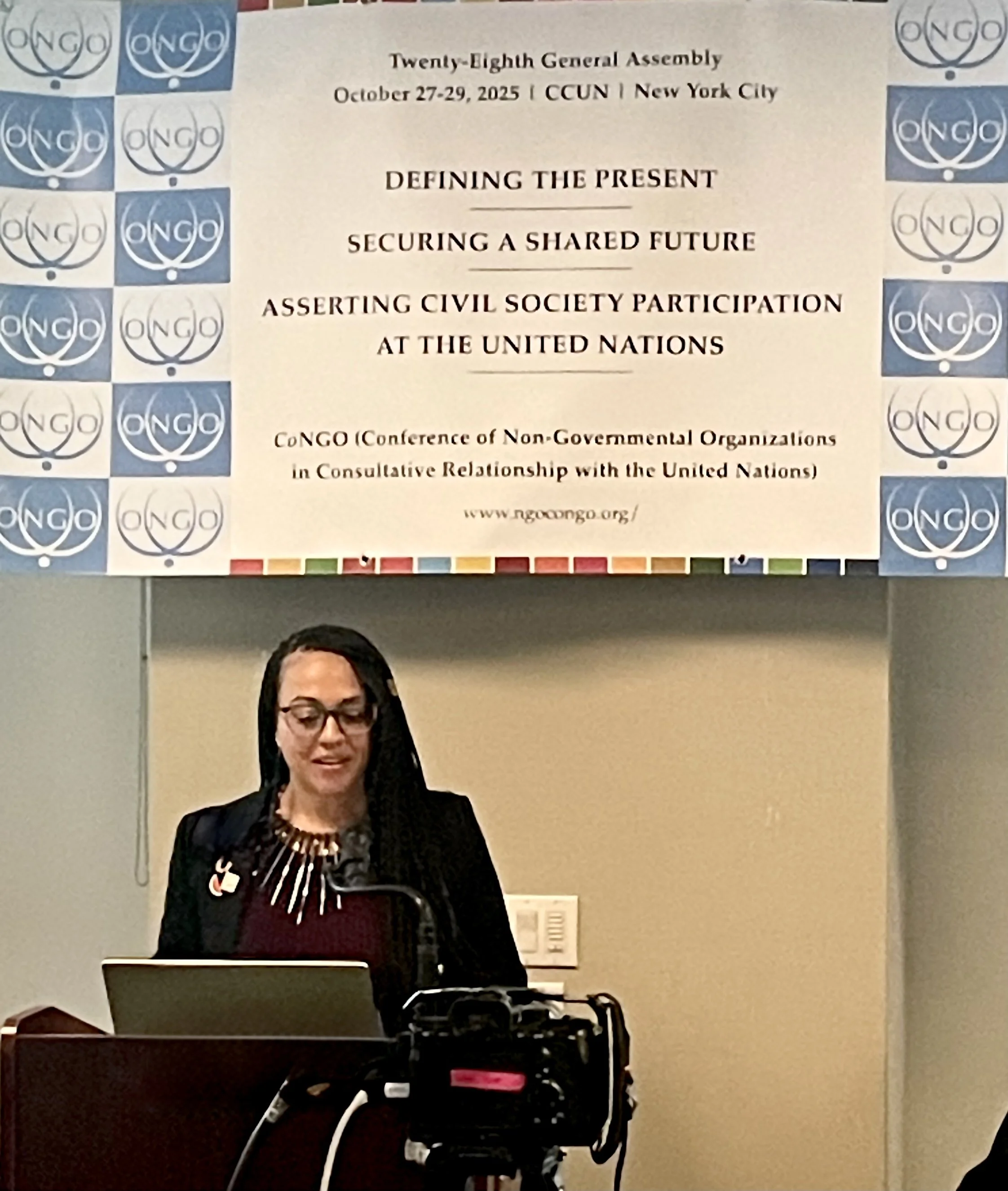 A woman giving a presentation at a conference with a large banner behind her that reads 'Twenty-Eighth General Assembly October 27-29, 2025 | CCUN | New York City' and 'Defining the Present, Securing a Shared Future, Asserting Civil Society Participa