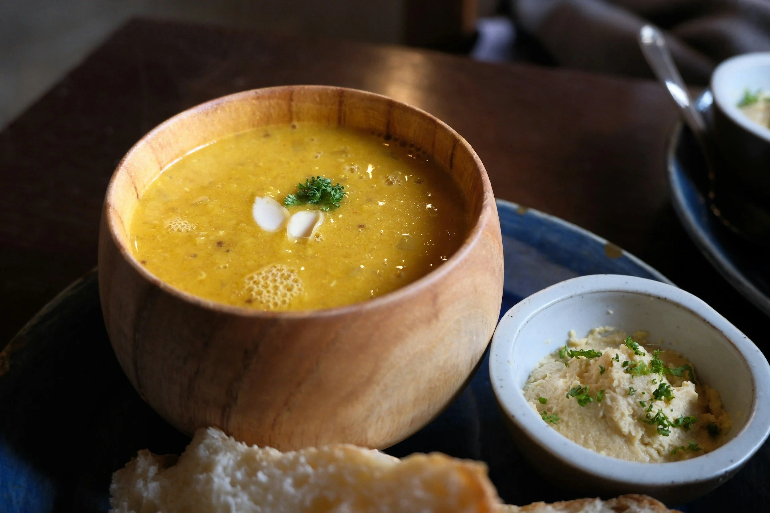 A bowl of yellow lentil soup garnished with parsley and sliced onions, served alongside a small bowl of mashed potatoes with herbs, and a piece of bread, all on a dark tray.