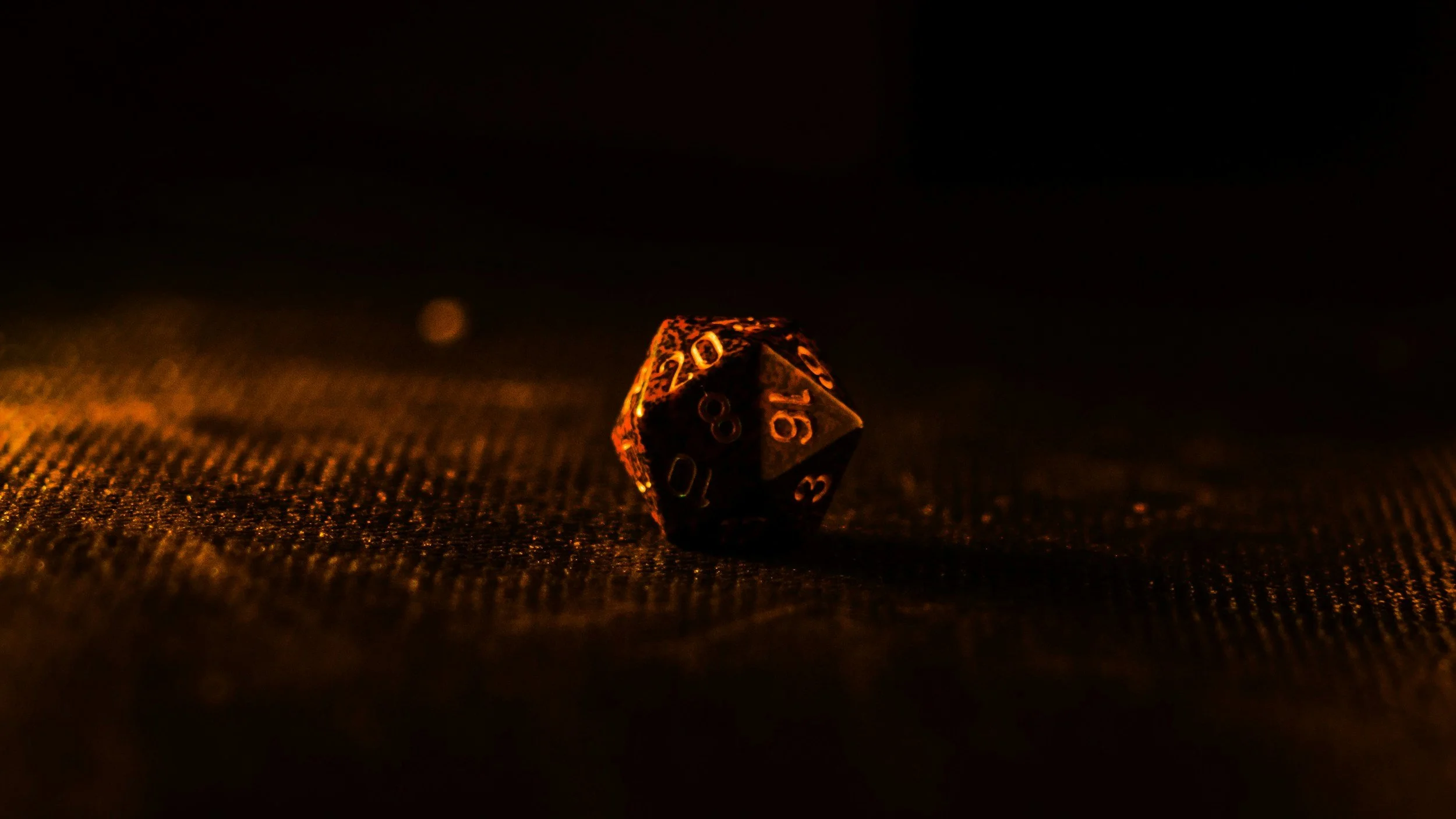 A metallic twenty-sided die (d20) with numbers visible, illuminated in warm orange light on a dark textured surface.
