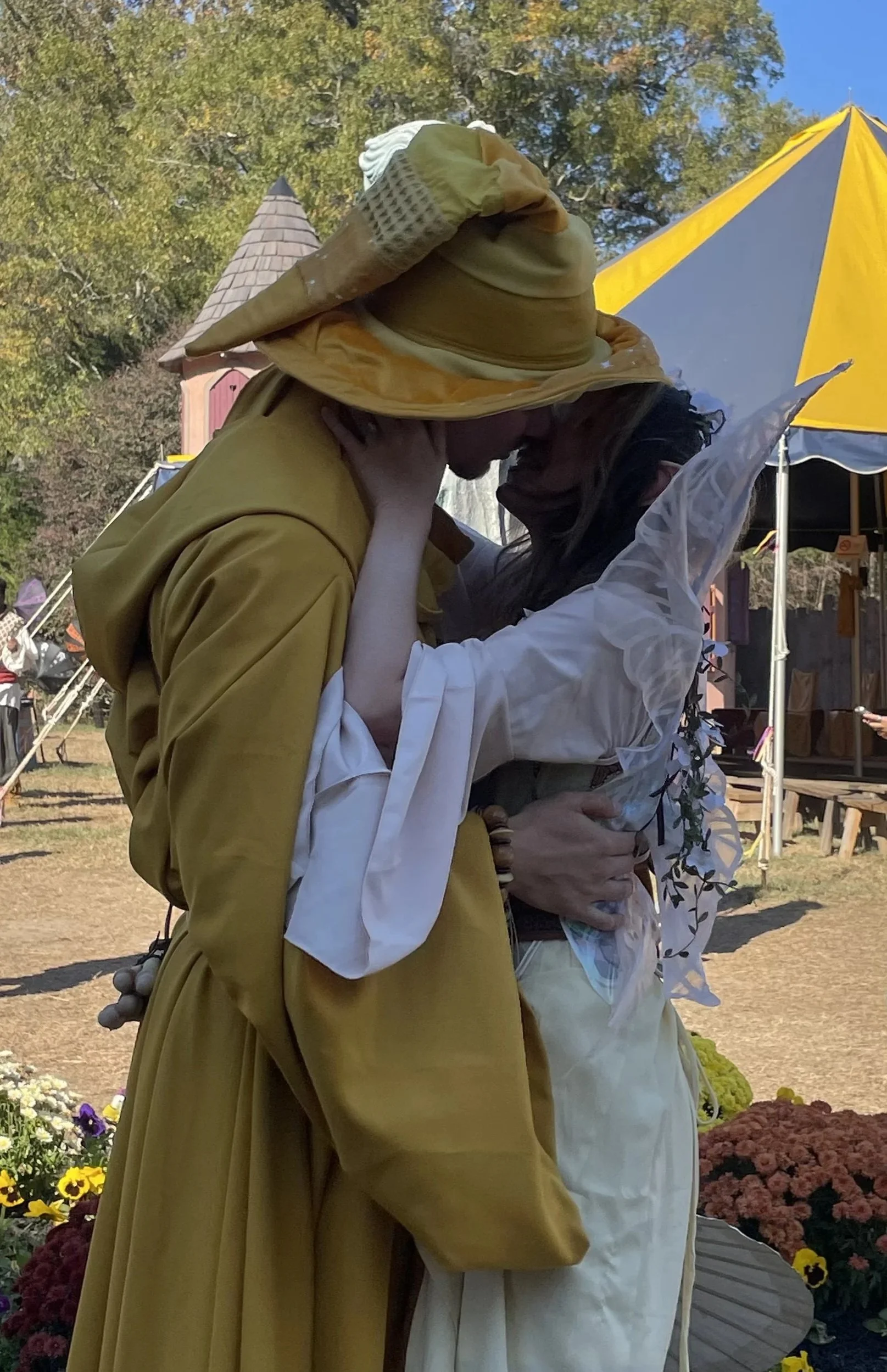 Two people dressed in medieval or fantasy costumes sharing a kiss at an outdoor event with yellow and blue tents and flowers in the background.