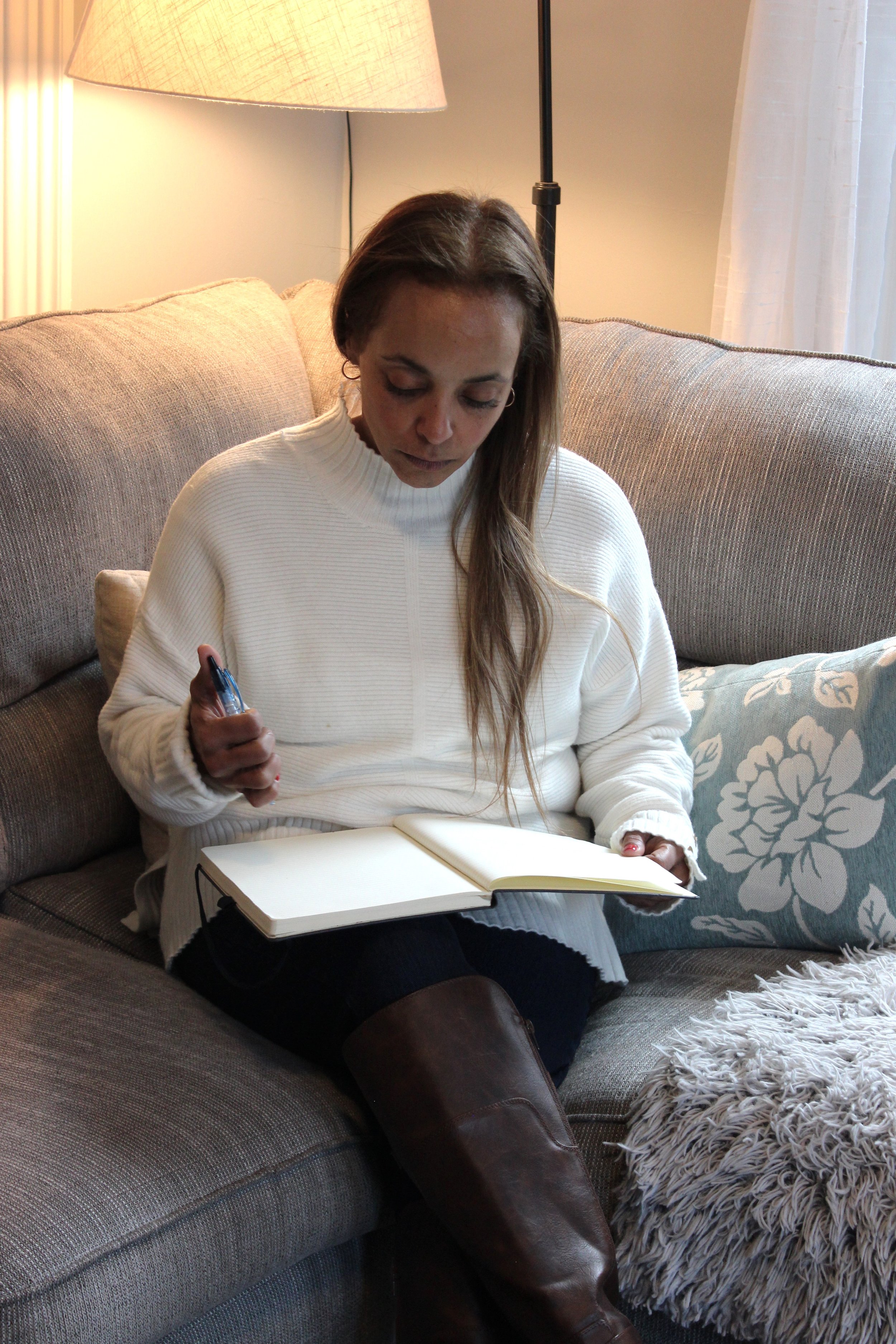 A woman sitting on a couch, looking down at a notebook, holding a pen in her left hand, with a lamp behind her to the left and a pillow with a floral pattern to her right.