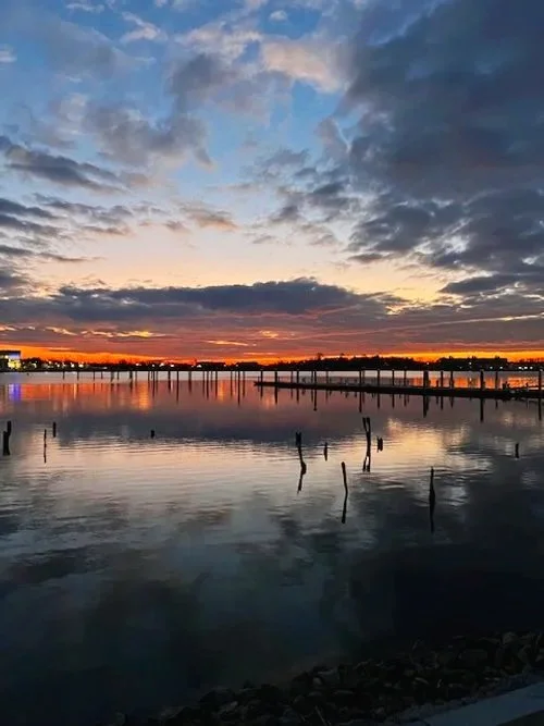 Sunset over a water body with cloudy sky and reflections of the sunset on the water, wooden posts in the water, and distant shoreline.