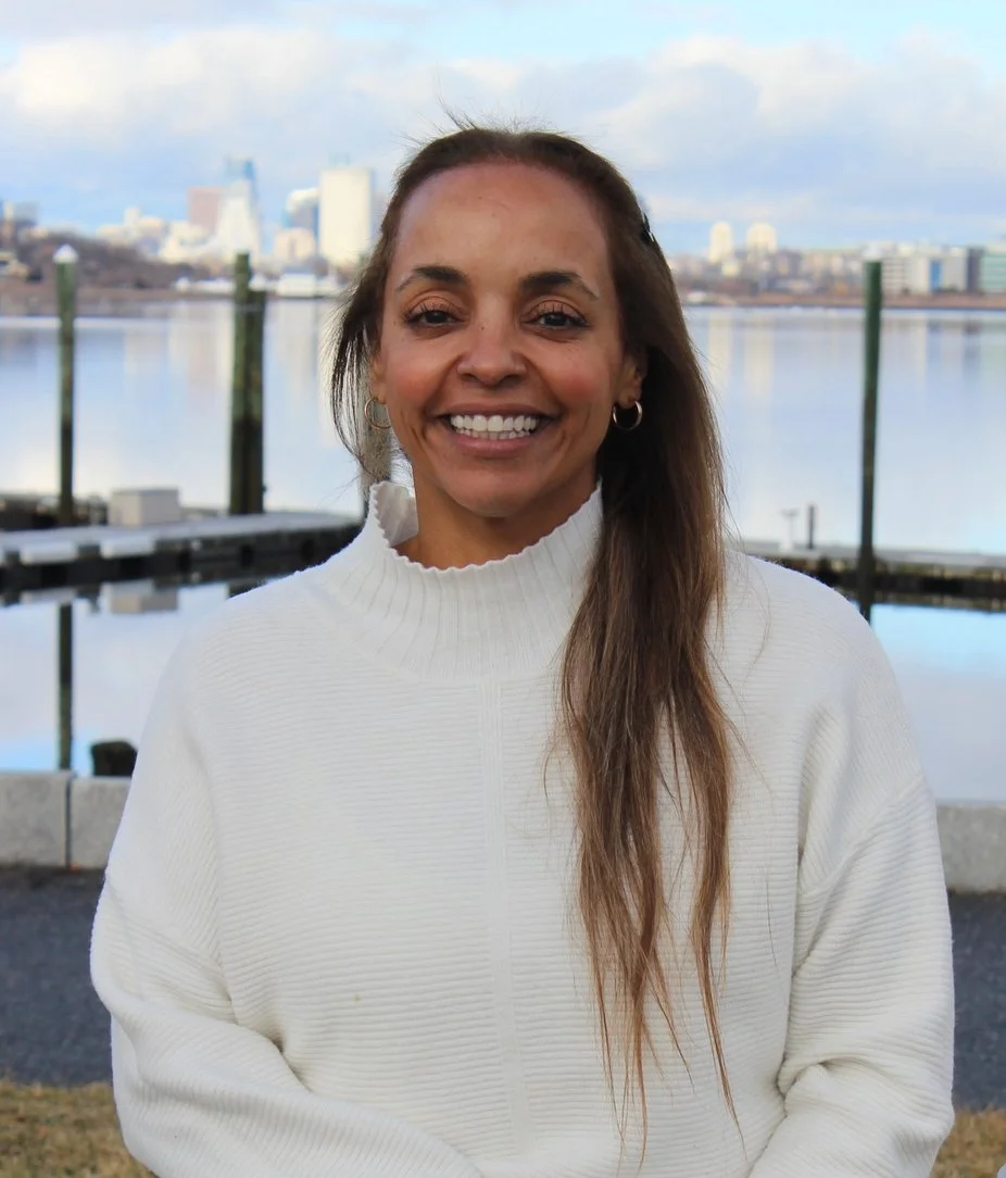 A woman with long brown hair smiling outdoors near a body of water with a city skyline in the background.