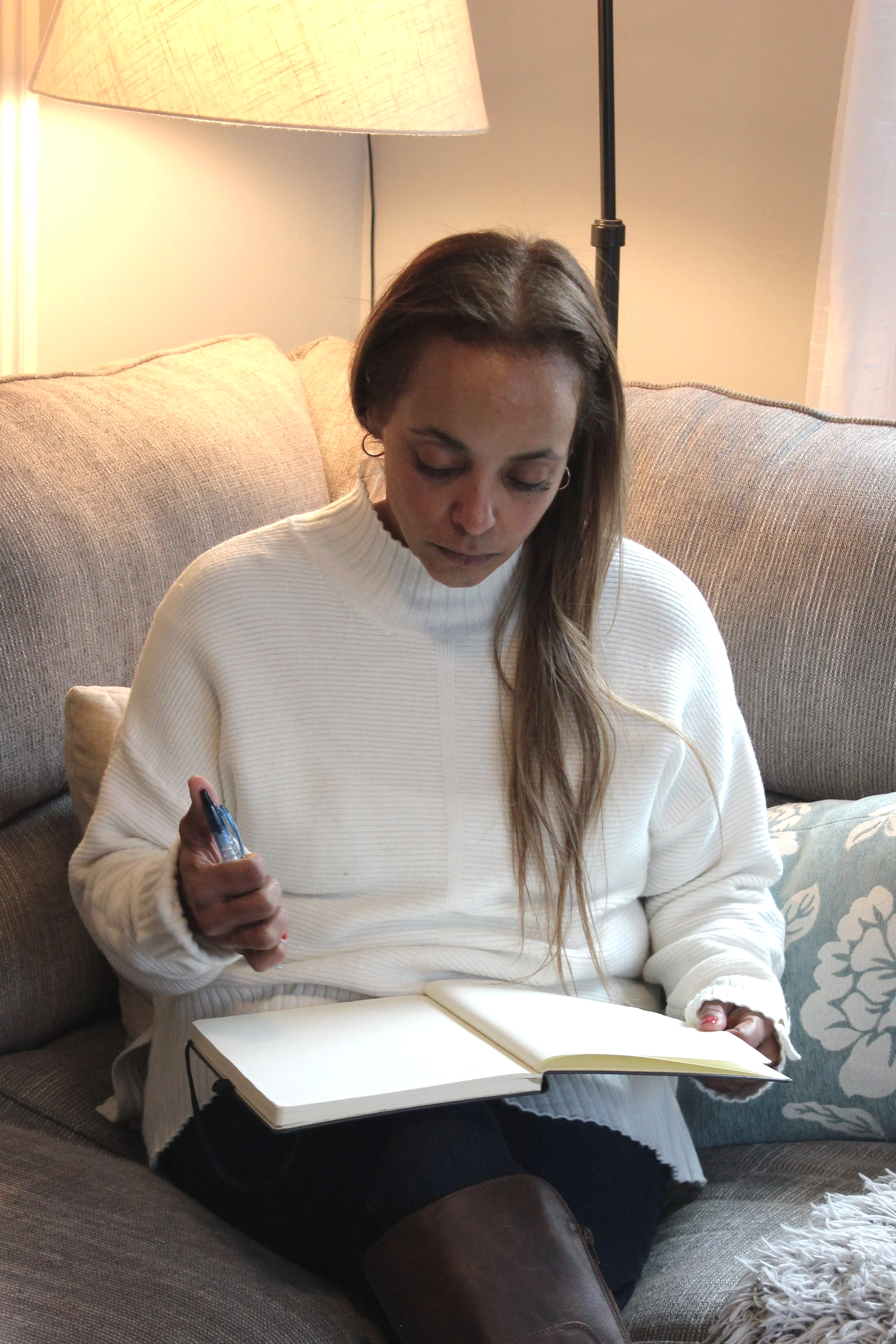 A woman sitting on a beige couch, looking down at an open notebook on her lap, with a pen in her right hand, in a cozy living room.