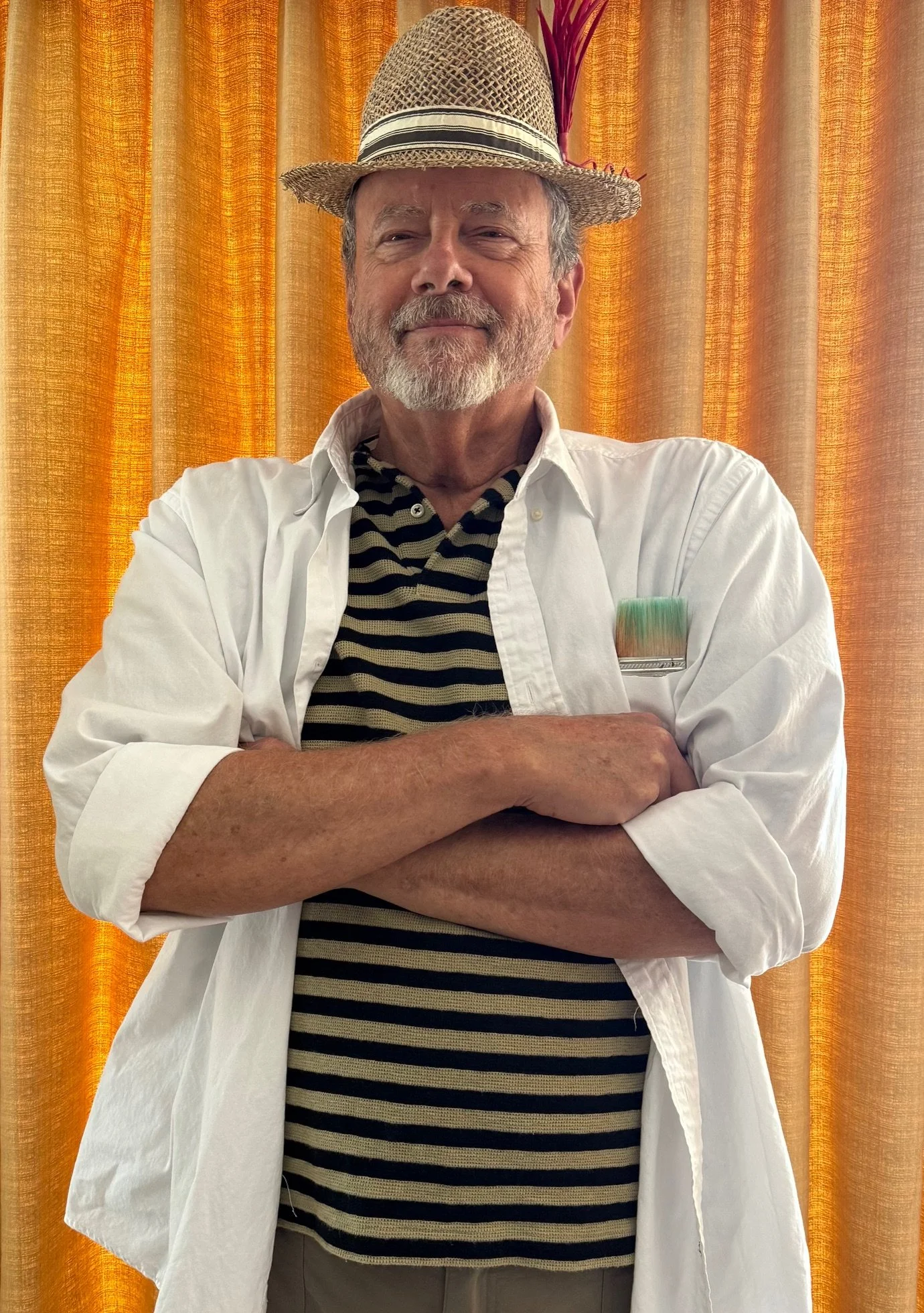 A man with a beard and mustache wearing a straw hat with a feather, a white open shirt over a striped black and beige shirt, and beige pants stands with arms crossed in front of orange curtains.