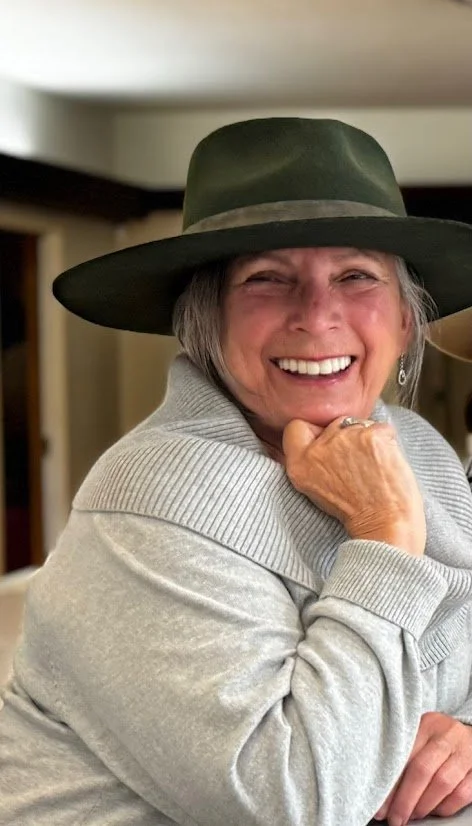 Smiling elderly woman wearing a gray sweater and a wide-brimmed hat.