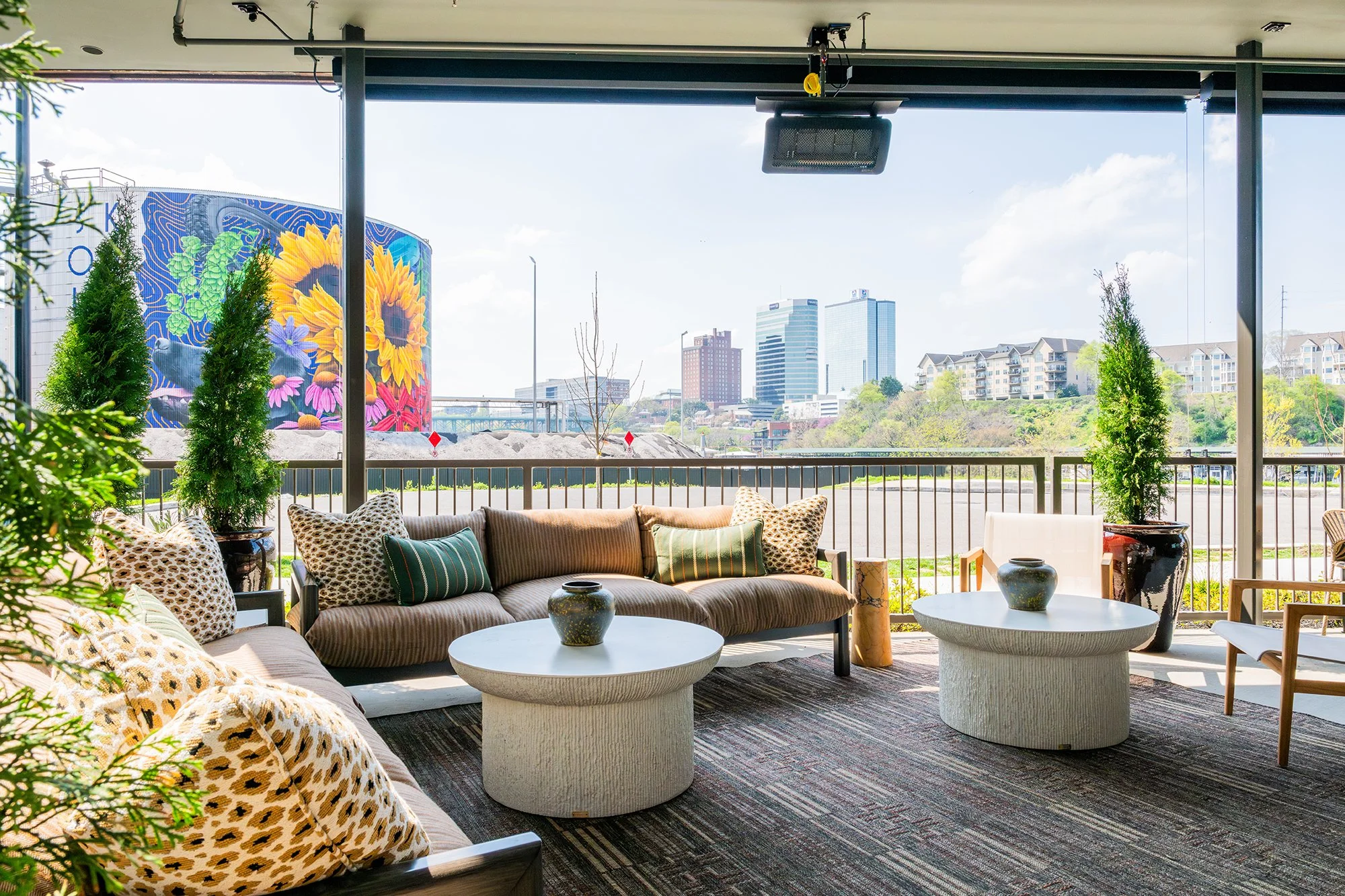 Outdoor balcony seating with patterned pillows and two white cone-shaped tables, overlooking a cityscape with tall buildings and a colorful mural with sunflowers and flowers.