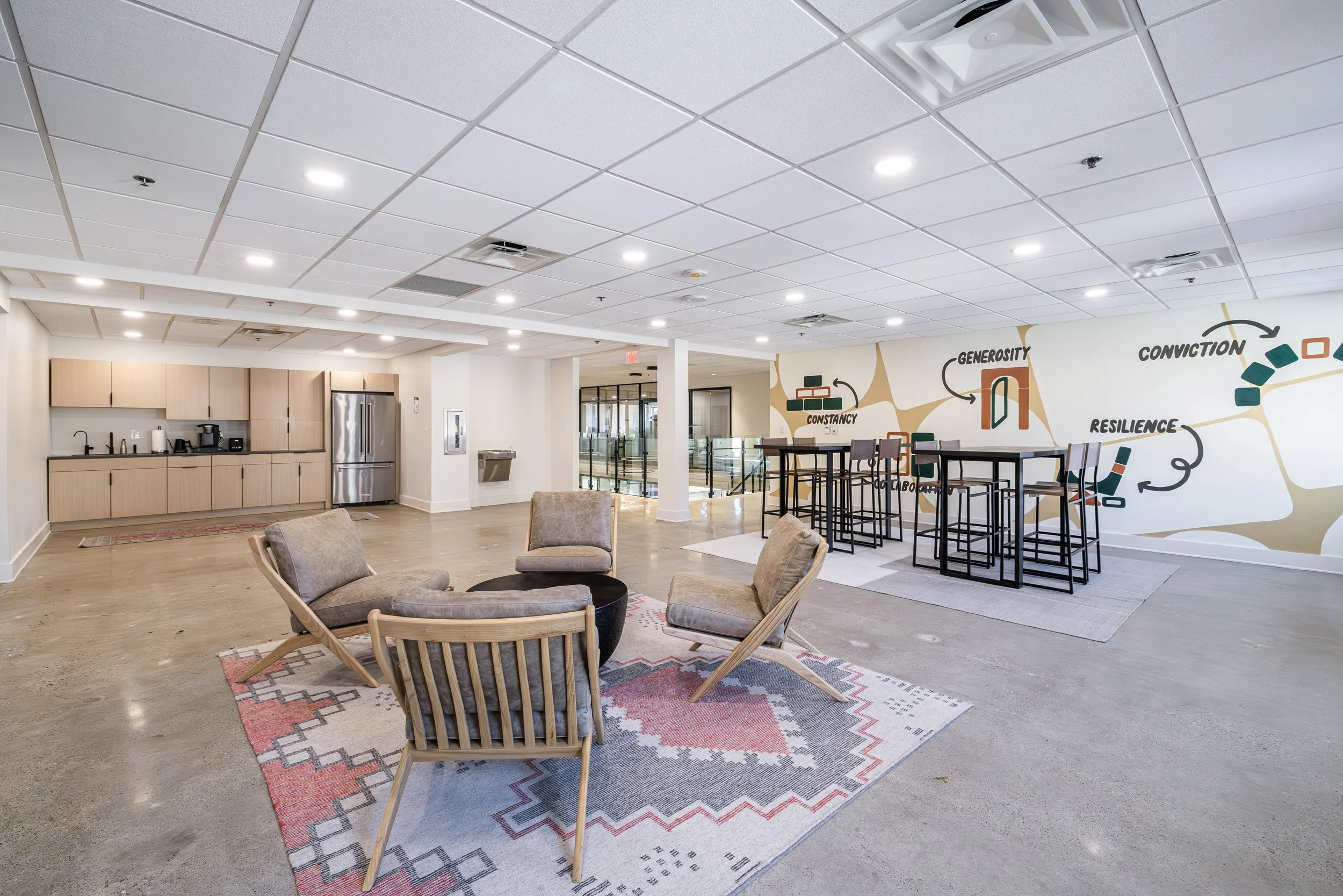 Modern office break room with wooden cabinets, stainless steel refrigerator, coffee station, and wall mural with words like 'Generosity', 'Conviction', 'Resilience', and 'Constancy'. Seating area includes chairs, armchairs, and a colorful rug.
