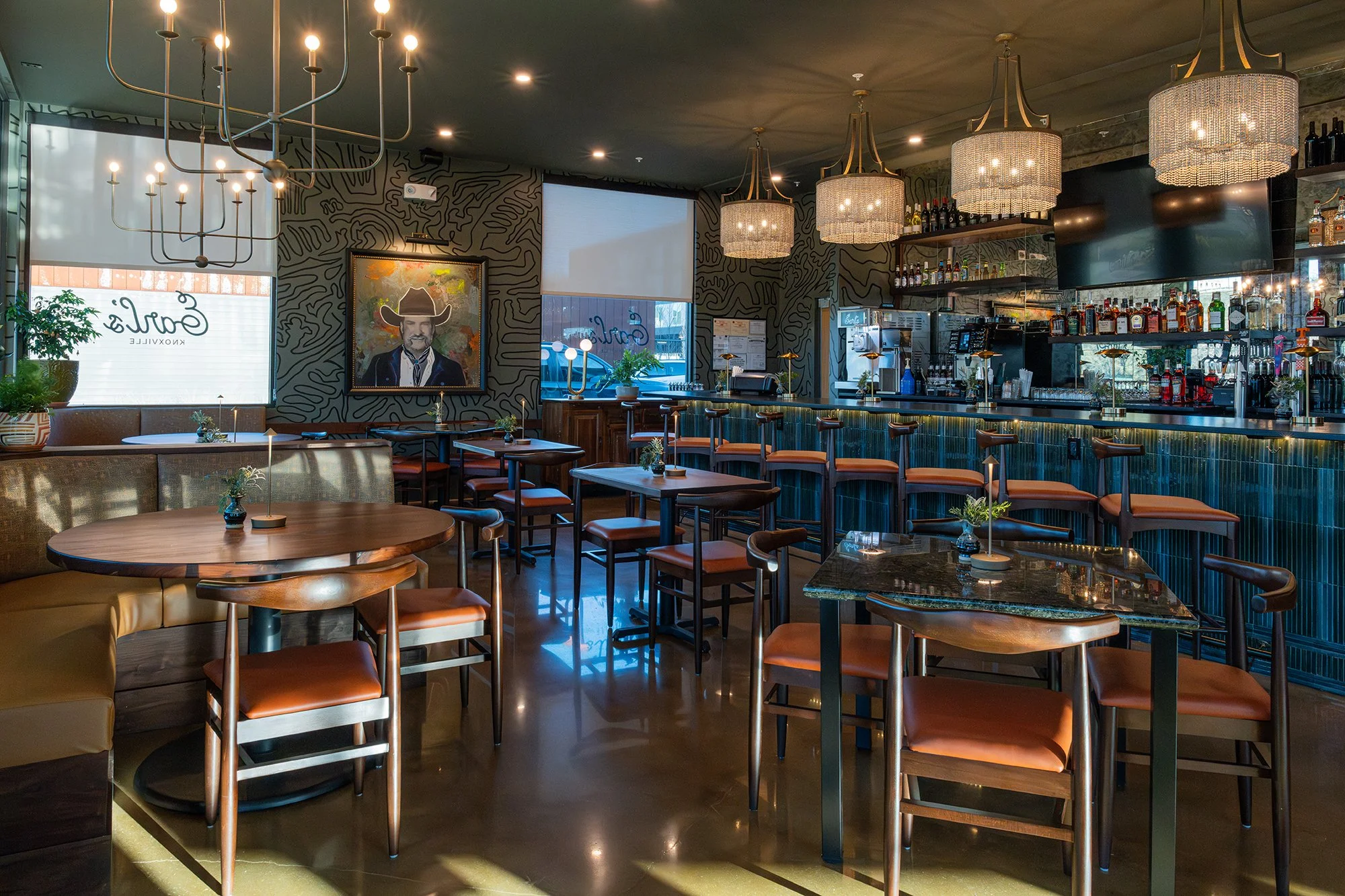 Interior of a stylish bar or restaurant with wooden tables and chairs, a bar counter with bottles of alcohol, large hanging chandeliers, a wall painting of a man with a cowboy hat, and a window with blinds letting in natural light.
