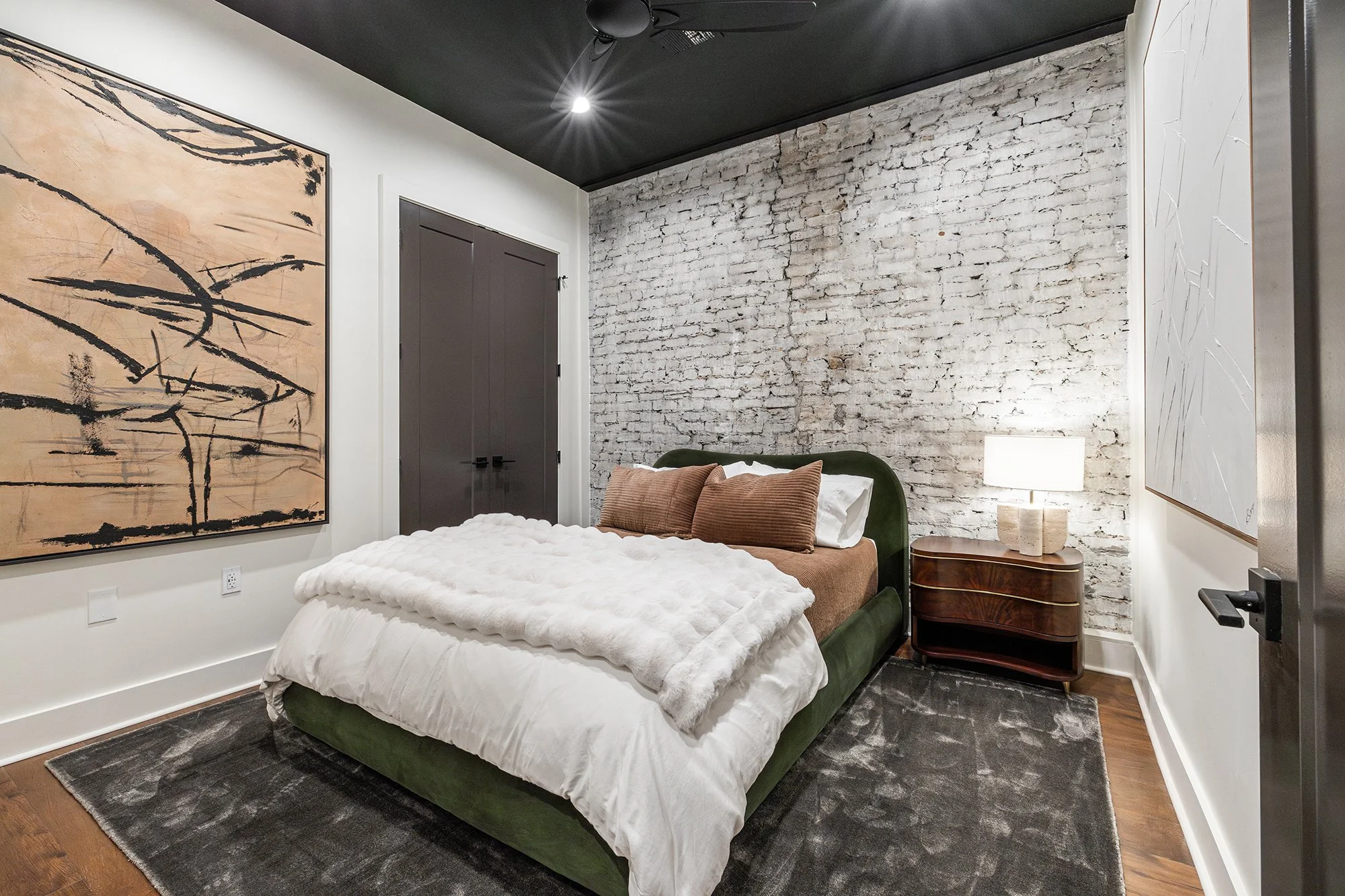 Bedroom with a green upholstered bed, brown pillows, exposed brick, and abstract artwork.