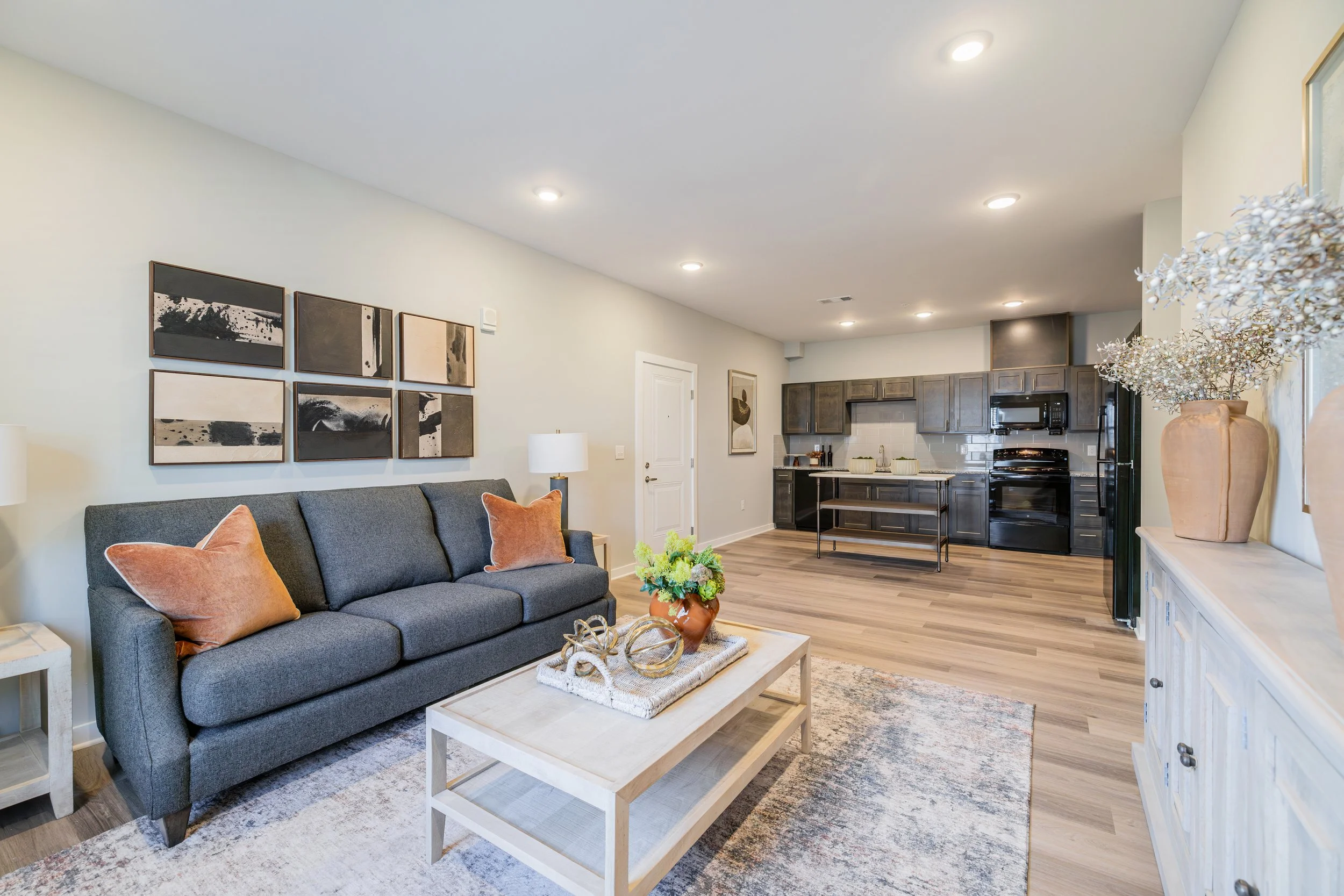Living room with a gray sofa, orange pillows, a wooden coffee table decorated with a flower vase, abstract wall art, and an open kitchen with dark cabinets and stainless steel appliances.