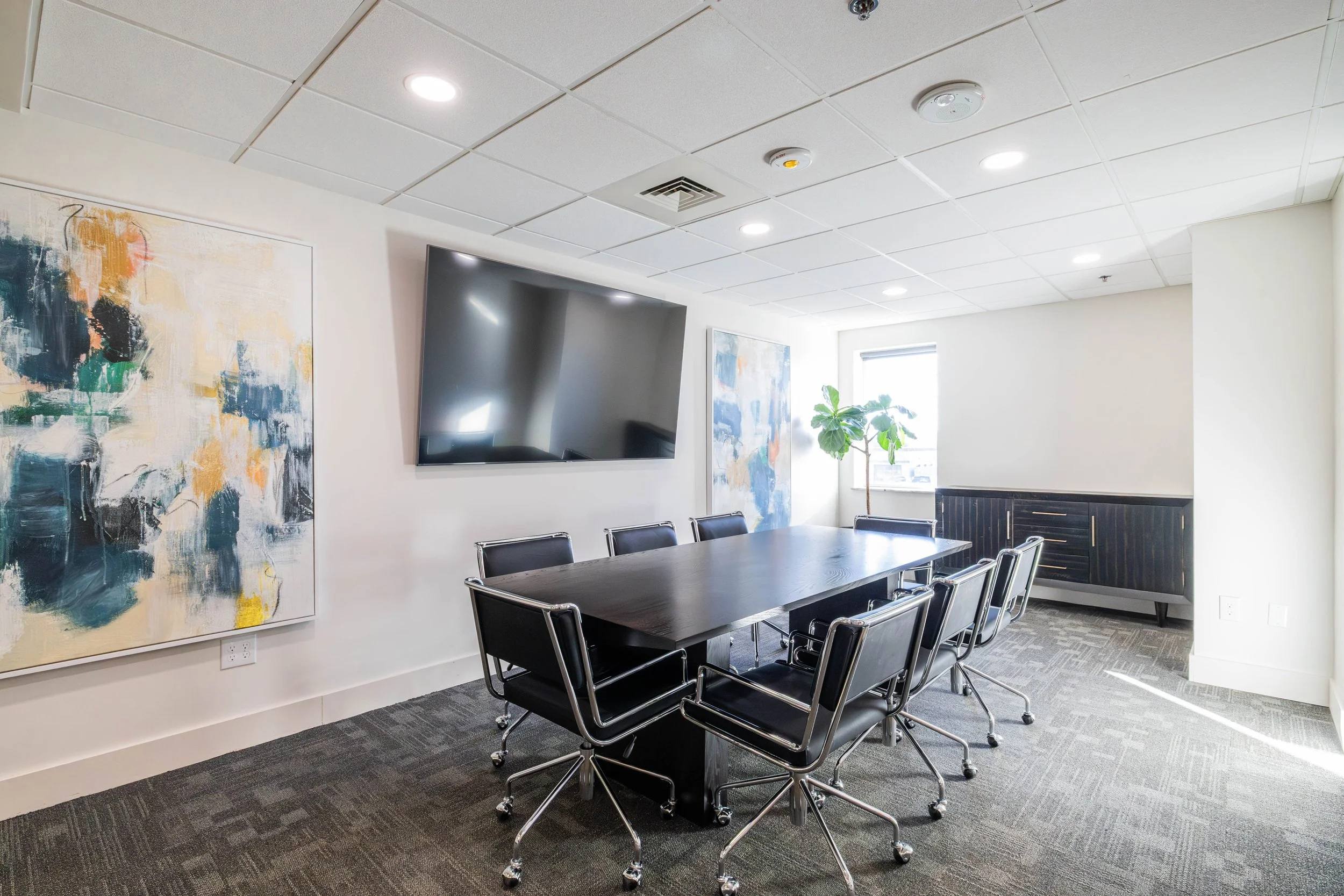 A modern conference room with a large black table surrounded by black chairs with chrome frames, abstract artwork on the white walls, a flat screen TV on one wall, a large window with blinds, and a potted plant near the window.