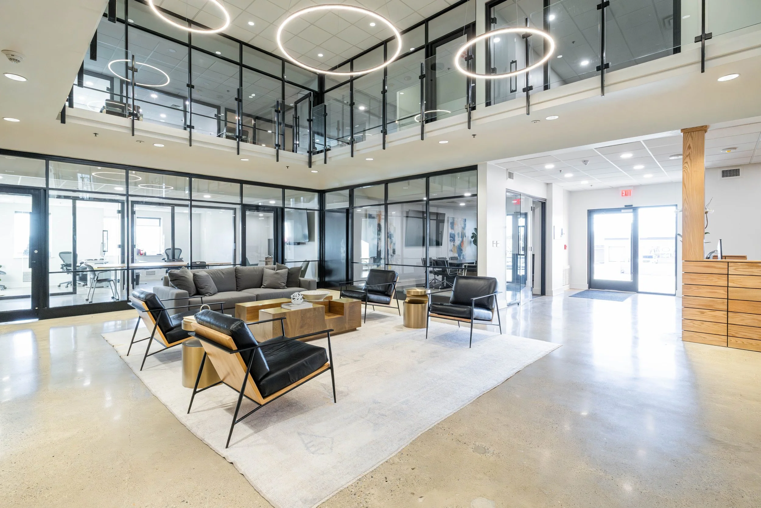 Modern office lobby with glass-walled meeting rooms, contemporary lighting, and stylish seating area with black chairs, a grey sofa, and a wooden coffee table.