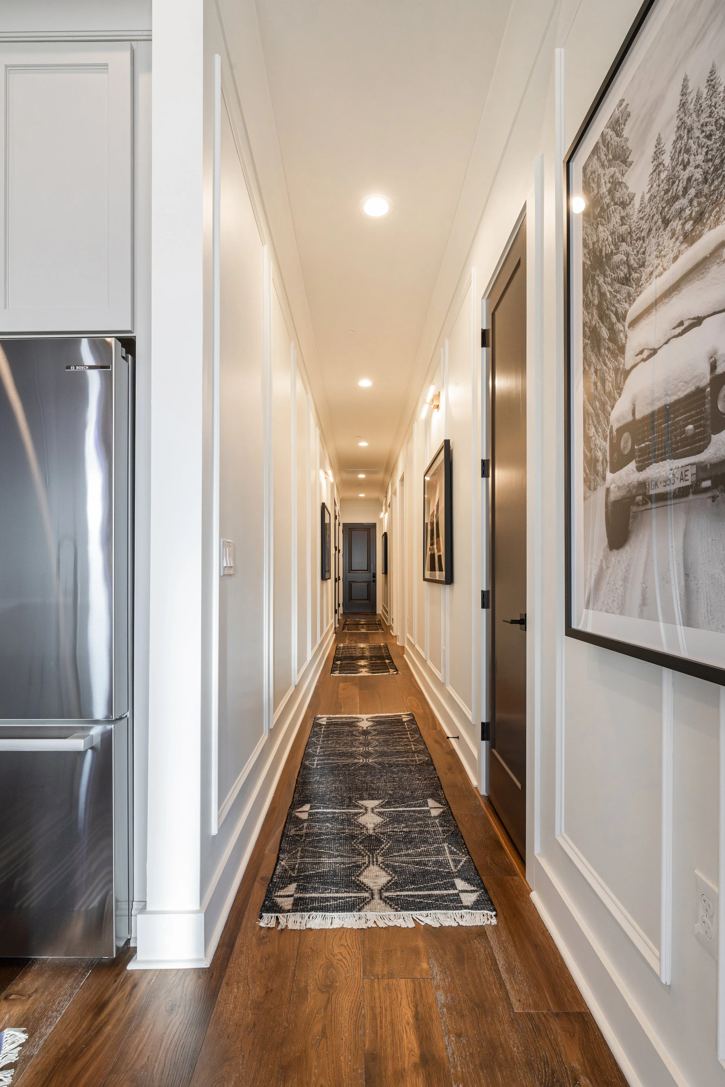 Long hallway with wooden floor, white walls, framed artwork, and patterned rug runners.