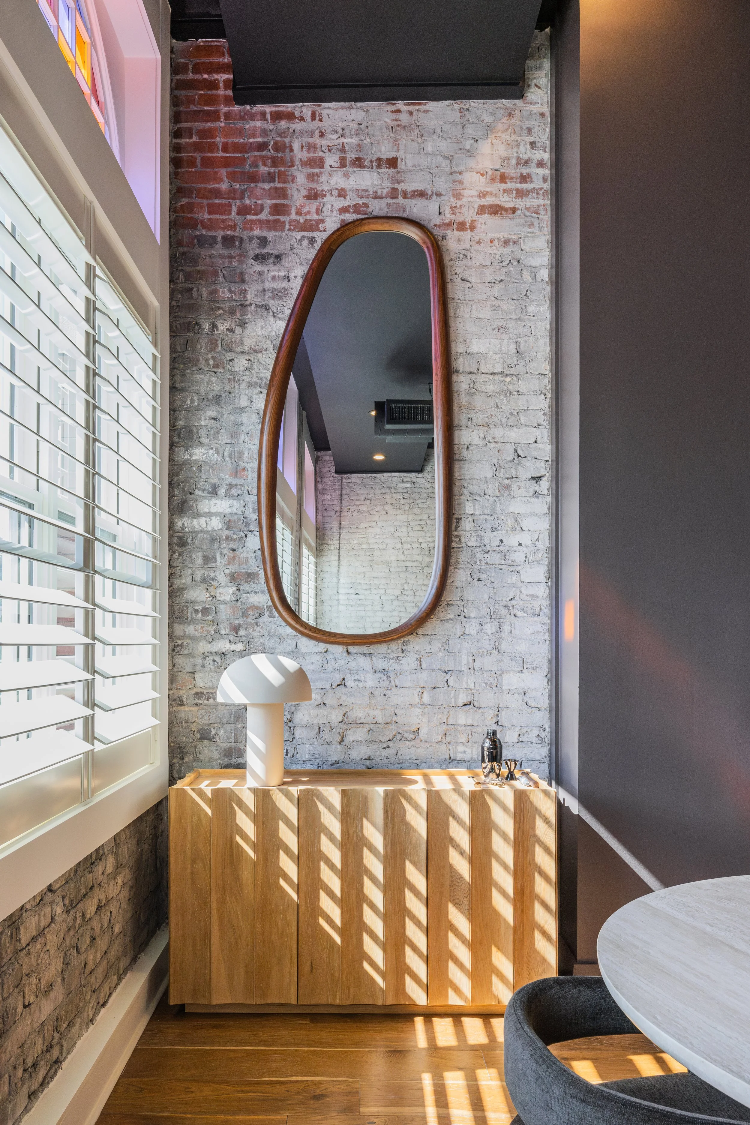 Sunlit corner with exposed white brick, a wood cabinet, and a tall arched mirror.