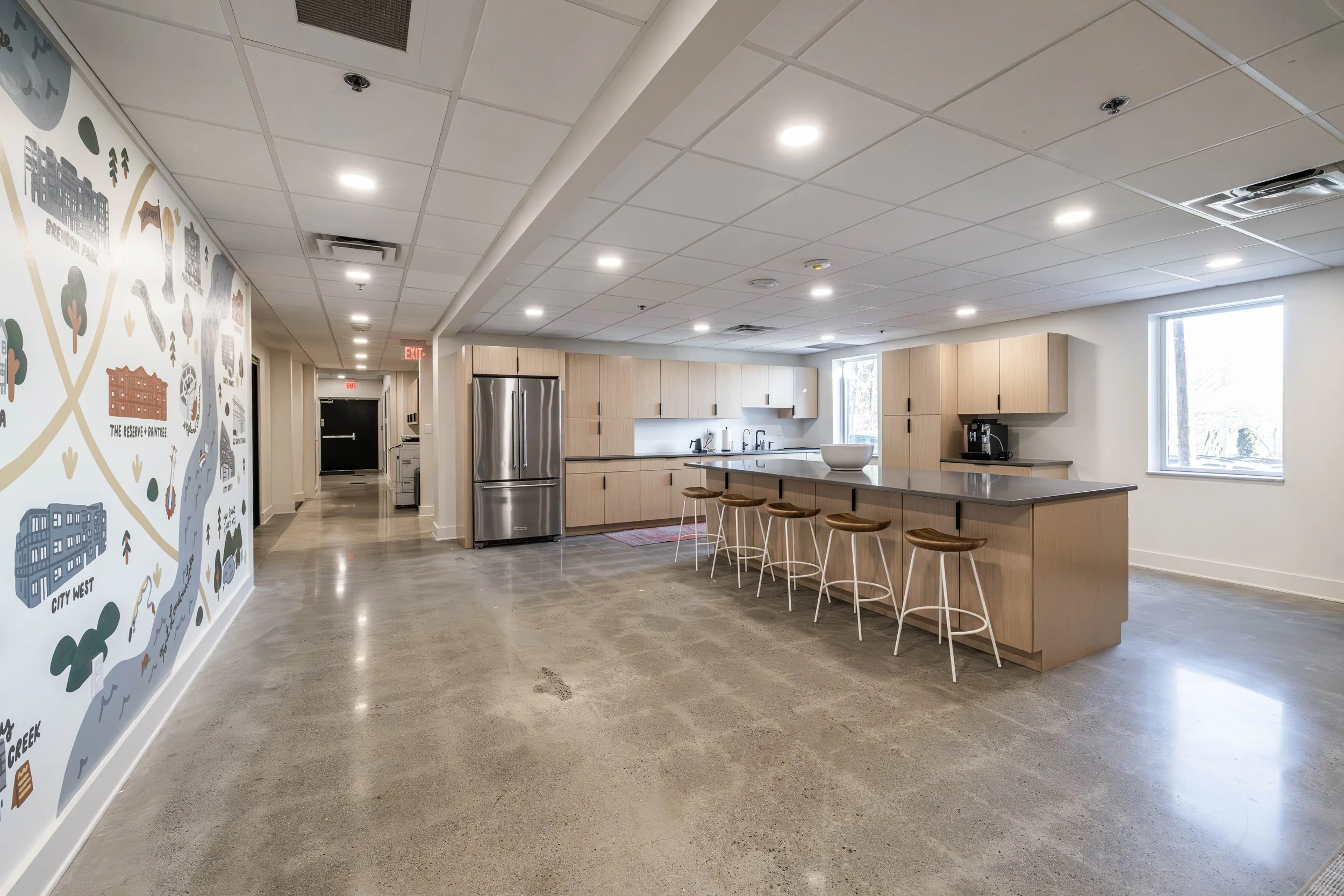 A modern kitchen area with light wood cabinets, a gray countertop, and bar stools. There is a stainless steel refrigerator and a coffee maker on the counter. Large windows bring in natural light, and a wall features a colorful illustrated map of a ci