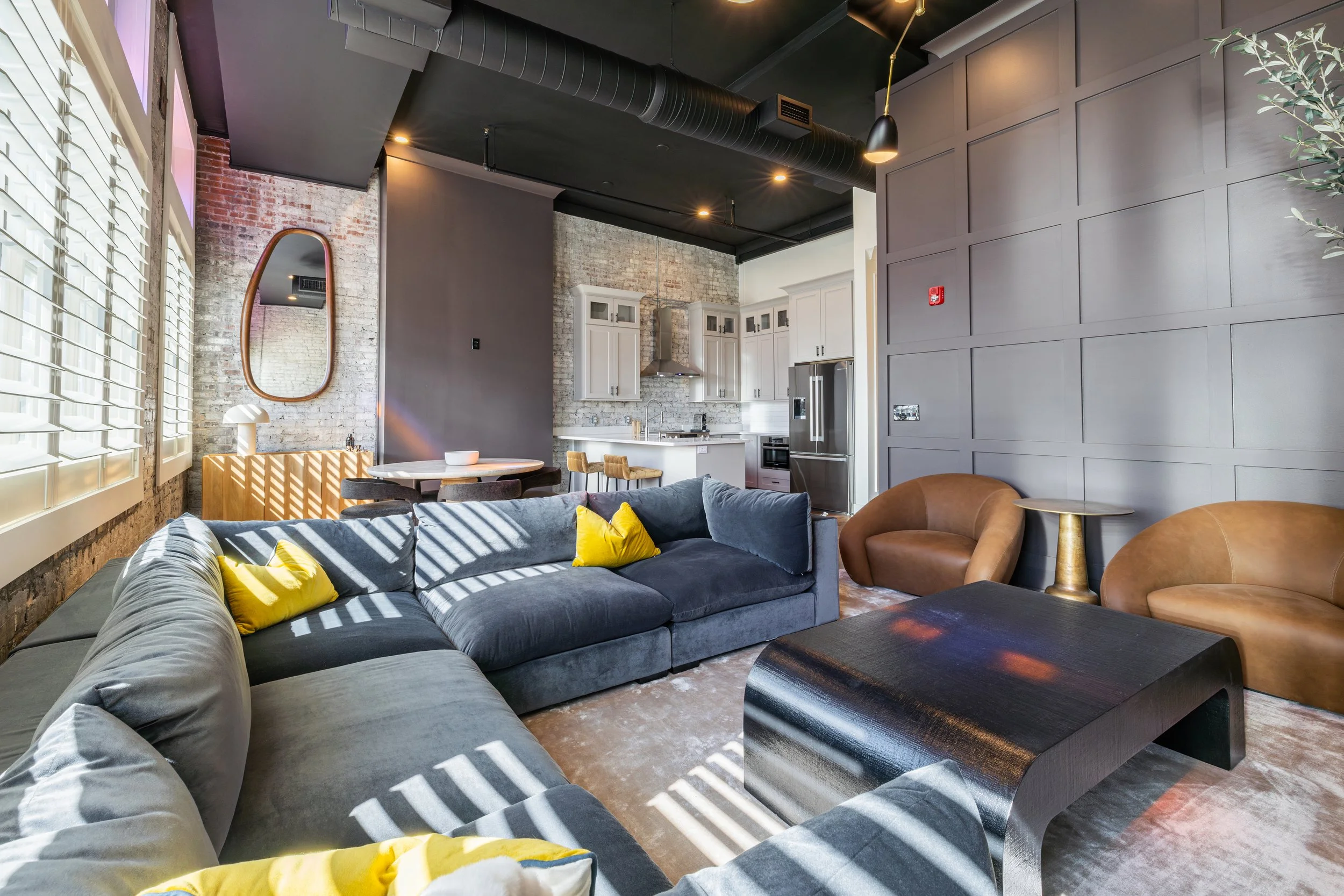 Sunlit lounge with a gray sectional, yellow pillows, exposed brick, and an open kitchen.