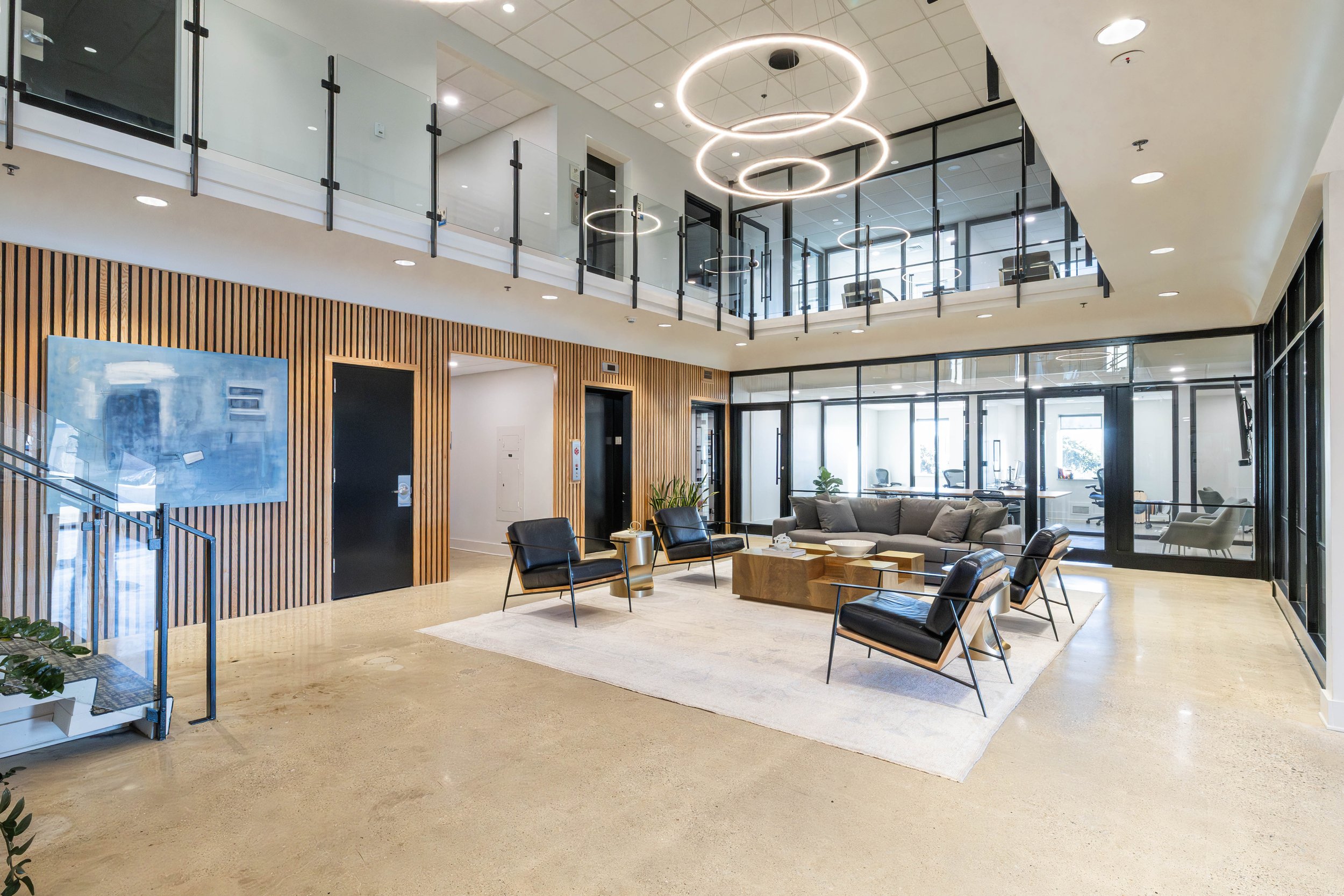 Modern office lobby with seating area, wooden panel walls, glass railing on an upper level, and large windows letting in natural light.
