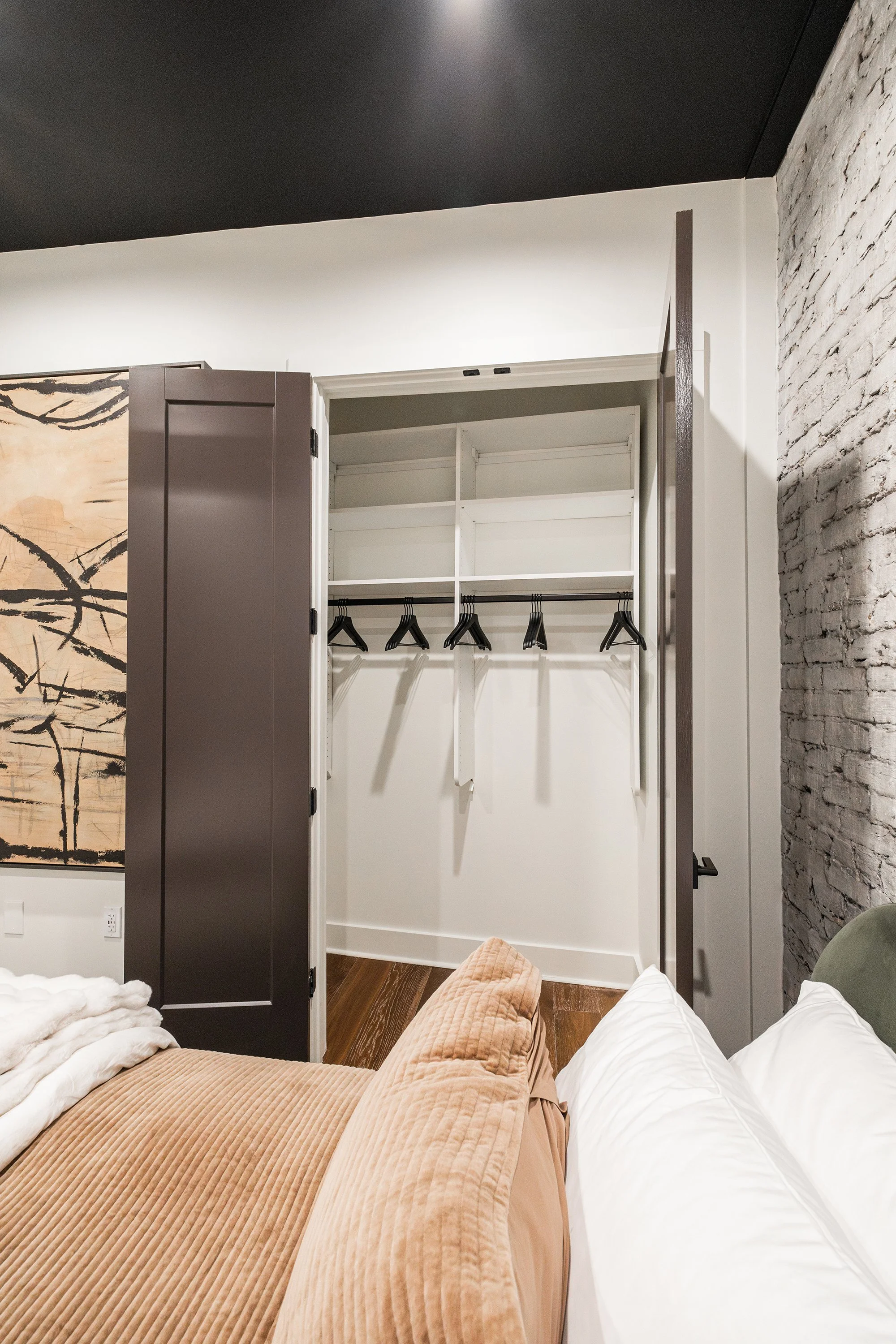 View of an open closet with white shelves, a hanging rod with black hangers, a beige corduroy armchair, white bedding, and artwork on the wall in a modern bedroom.
