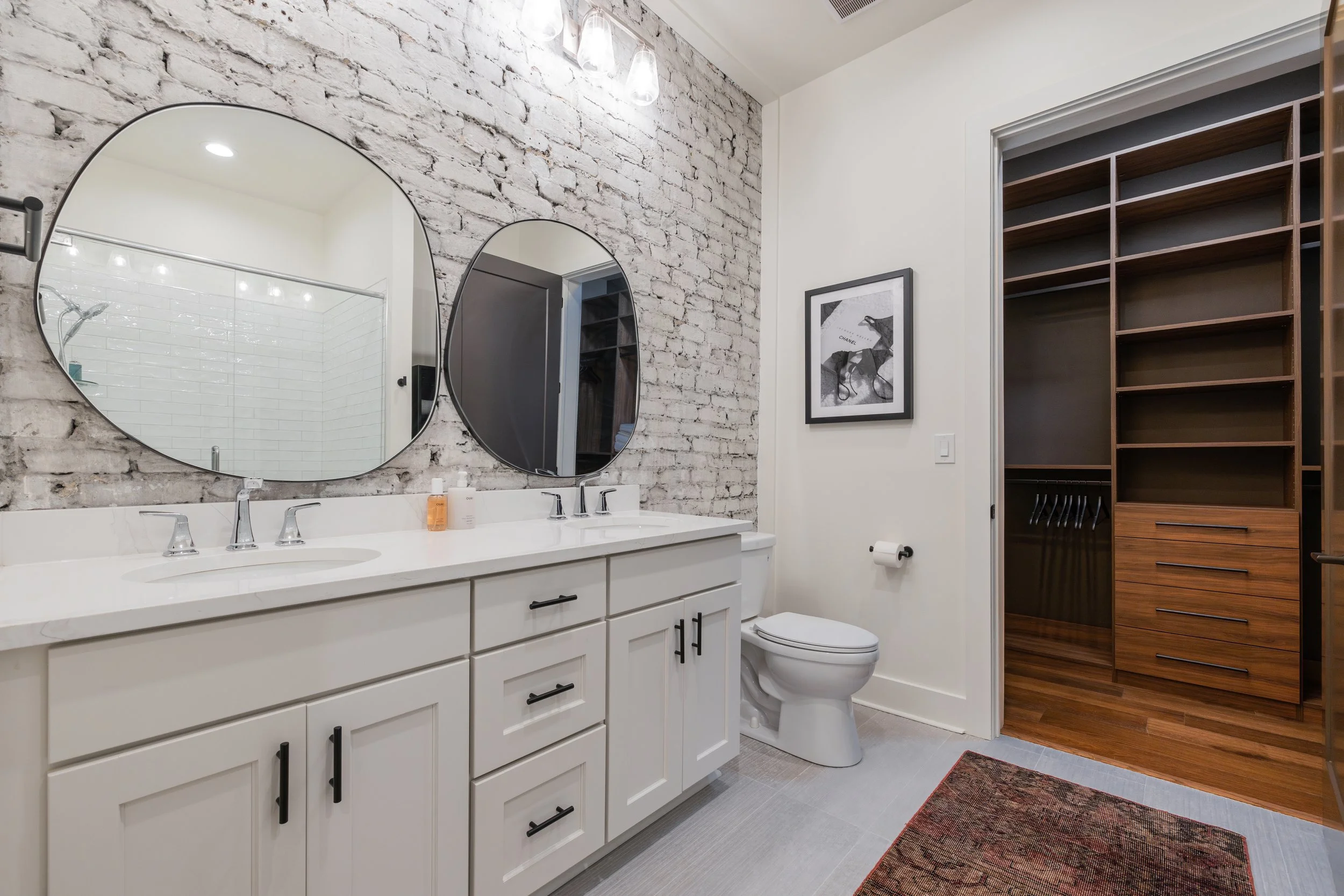 Bathroom with a double vanity, round mirrors, white brick, and an adjoining wood closet.