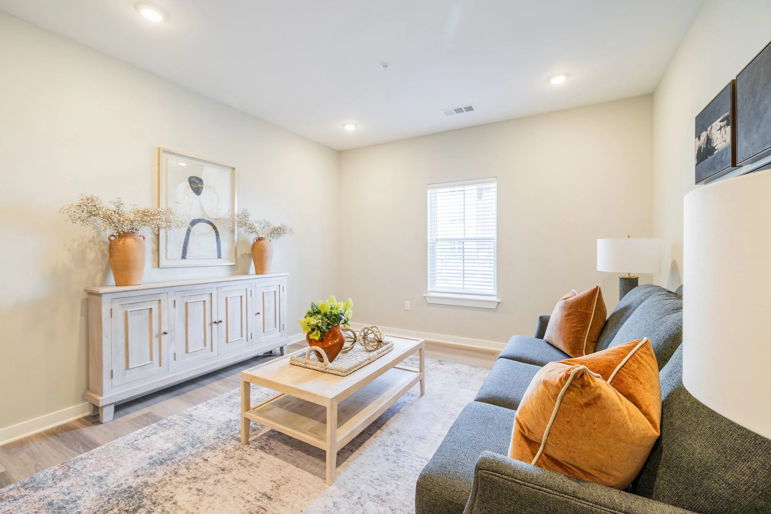 A cozy living room with a white sideboard, beige vases with dried flowers, a framed artwork, a window with blinds, and a gray sofa with orange and beige pillows. A wooden coffee table with a vase of yellow flowers is in front of the sofa. A beige rug