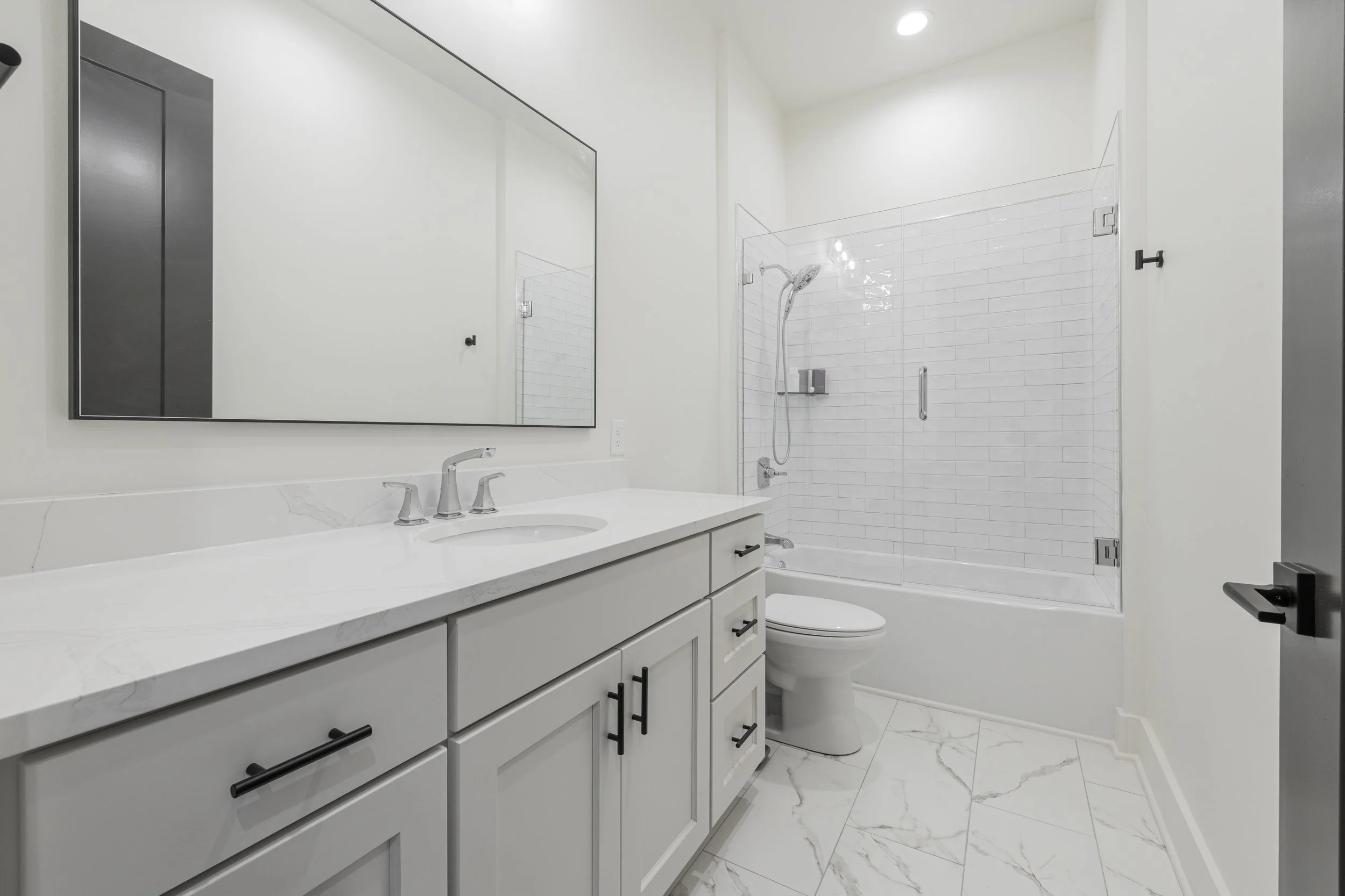 Modern bathroom with white vanity and marble countertop, large mirror, walk-in shower with glass door, white subway tiles, and a toilet.