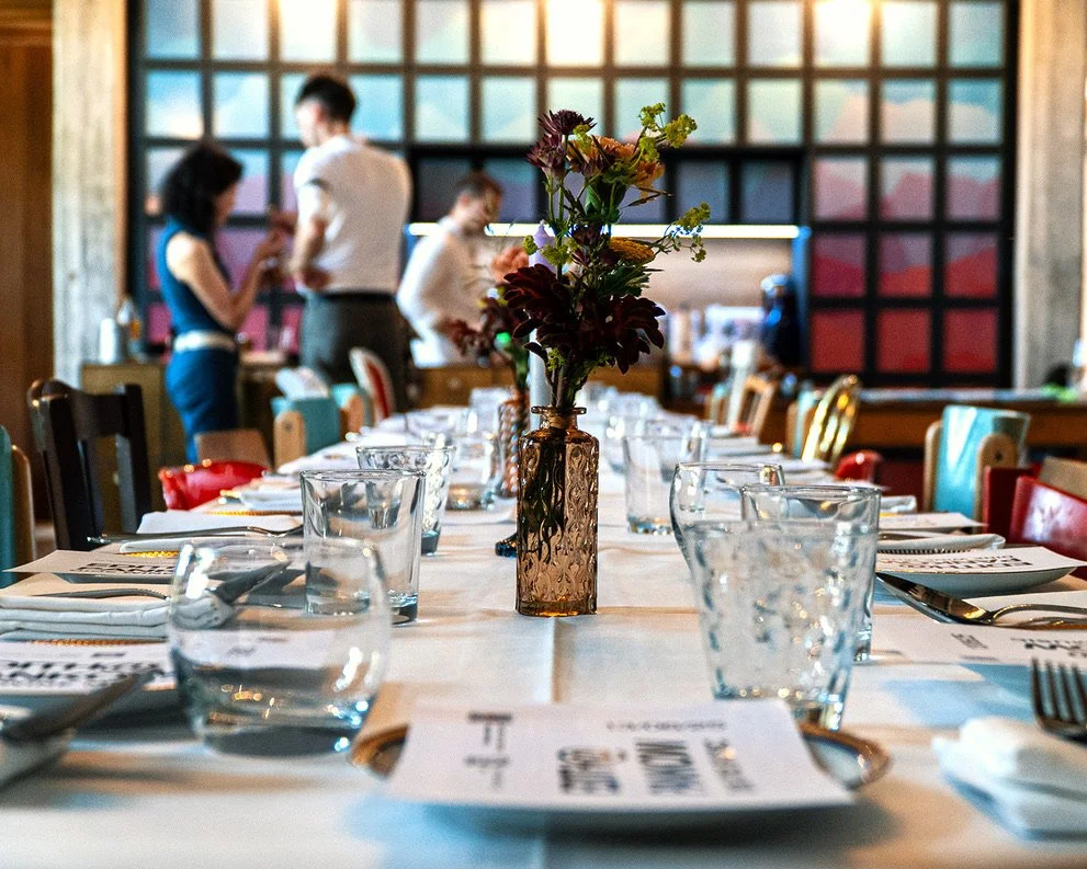 A long dining table set for a meal with glasses, plates, and a floral centerpiece in a restaurant with colorful wall art and blurred background of waitstaff.