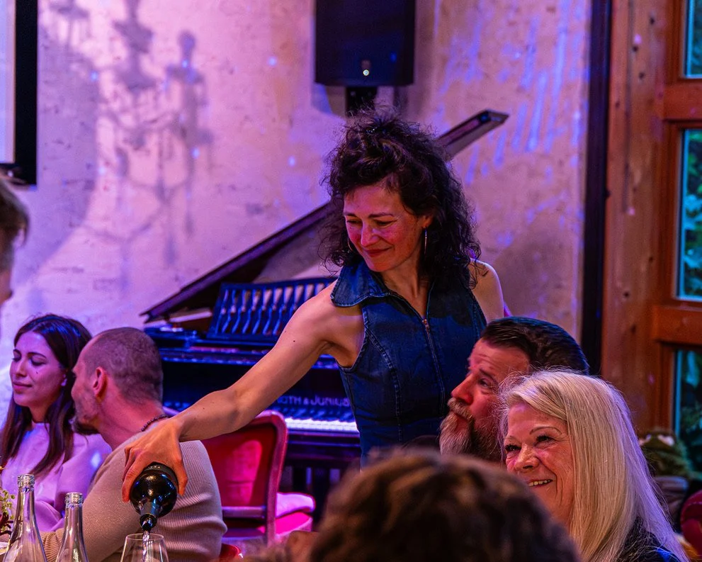 Woman in sleeveless denim top pouring drink at a social gathering indoors, with people seated at tables and a grand piano in the background.