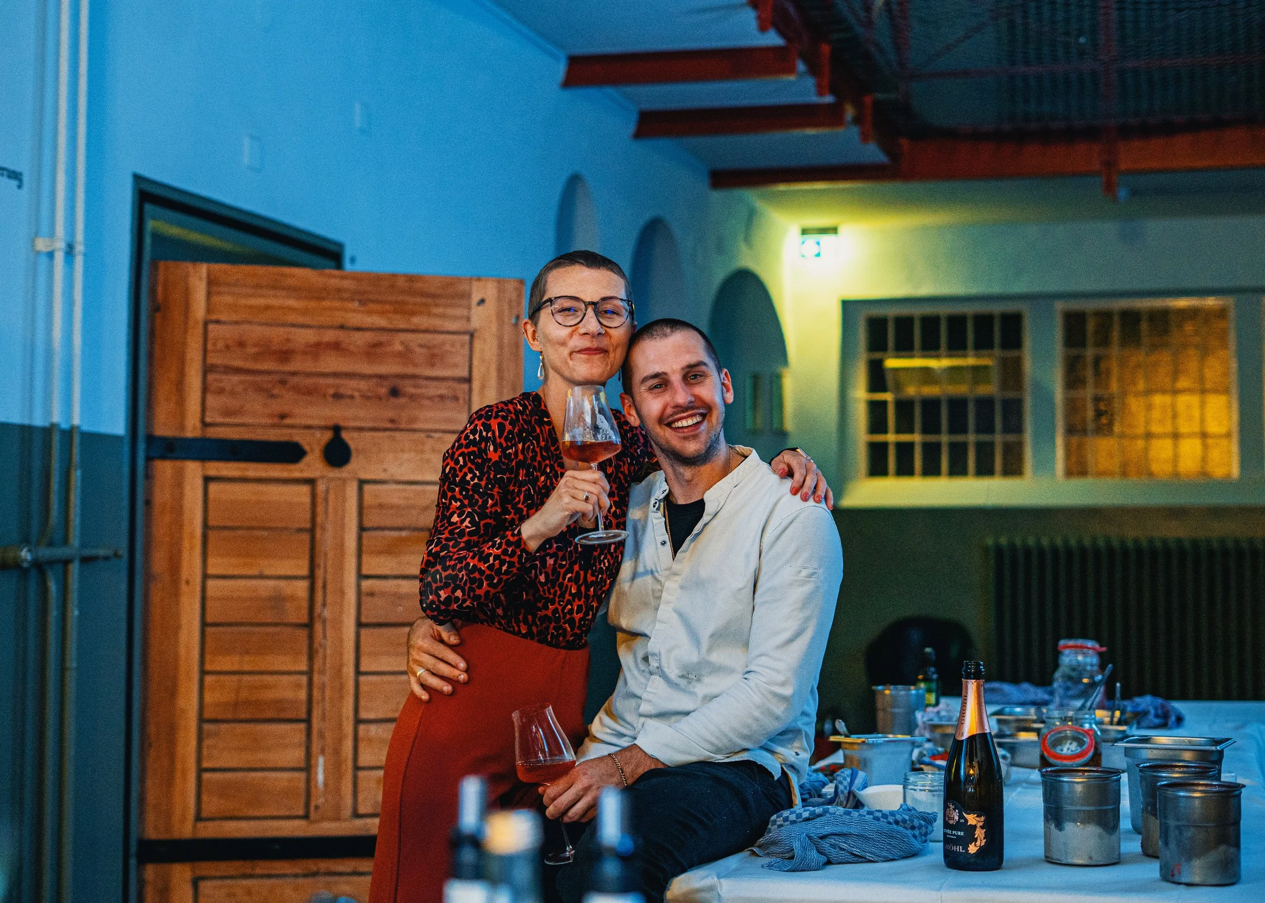 A woman and a man are smiling and posing together at a dinner party, with the woman holding a glass of wine and the man holding a glass of bubbly. They are inside a cozy, warmly lit room with decorated tables, bottles, and dinnerware around them.