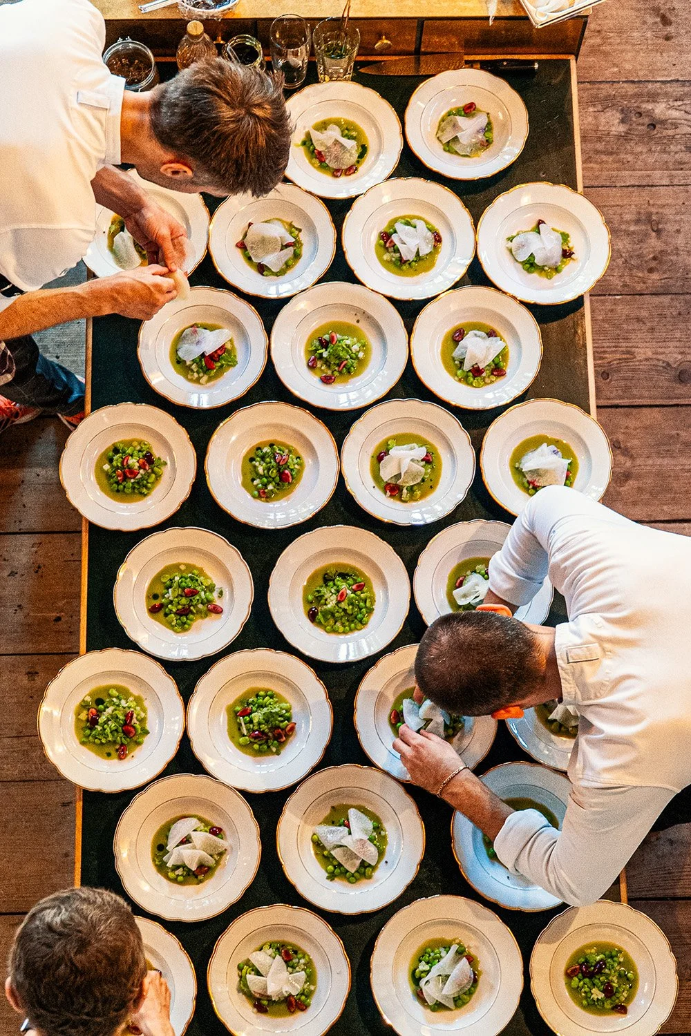Chefs preparing plates of green soup garnished with white cheese, pomegranate seeds, and fresh herbs on a long wooden table.