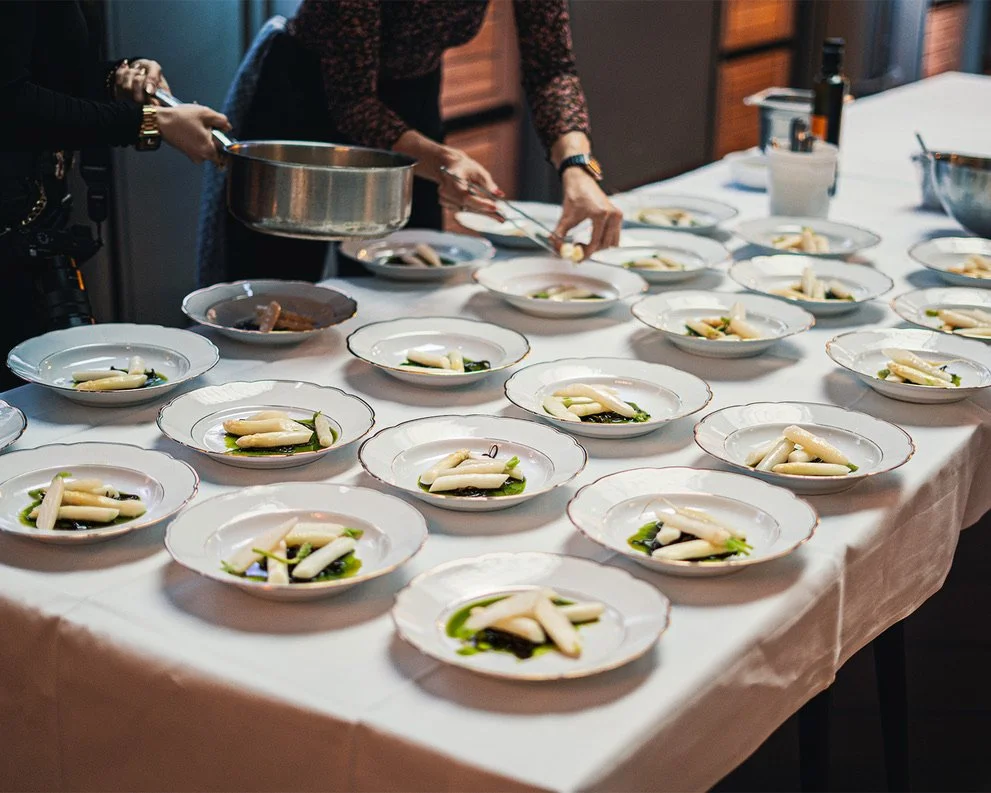 Multiple white plates with edamame and greens on a table, with chefs plating food in the background.